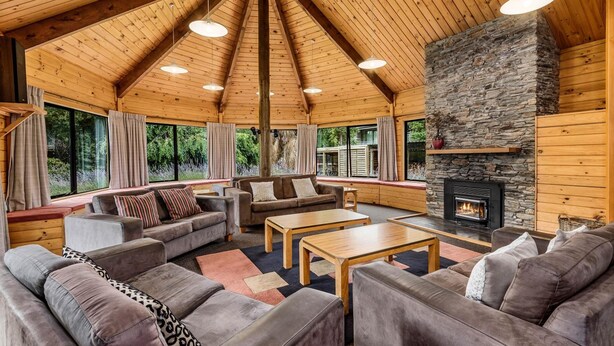 A cozy cabin interior with a stone fireplace, wooden ceiling, and large windows offering a view of the forest. - Altamont Lodge (Wanaka)