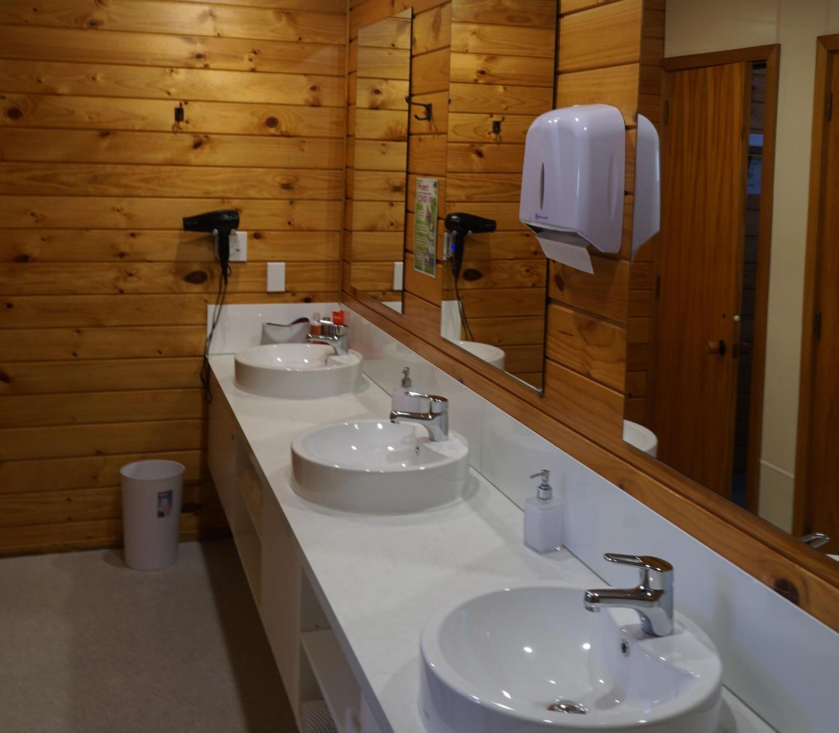 Une salle de bain de style rustique, avec des murs en bois, trois lavabos, un grand miroir et un sèche-cheveux.