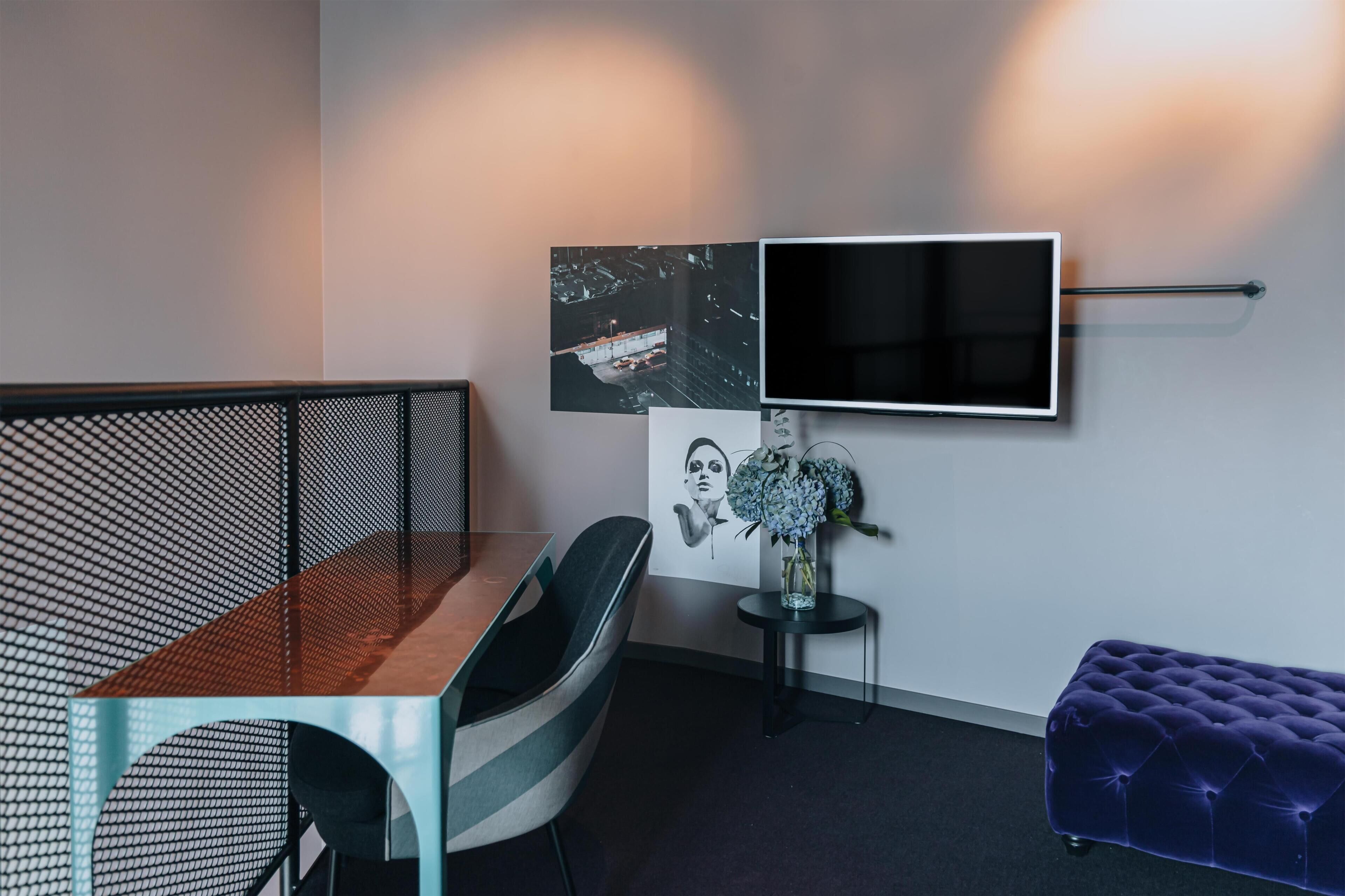 Quarto moderno com escrivaninha, cadeira, sofá, televisão, obras de arte e uma mesinha de centro com um vaso.