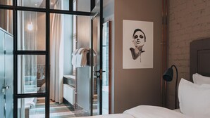 Une chambre d’hôtel avec un lit, une œuvre d’art accrochée au mur, une lampe et une vue sur la salle de bain, visible à travers une porte vitrée.