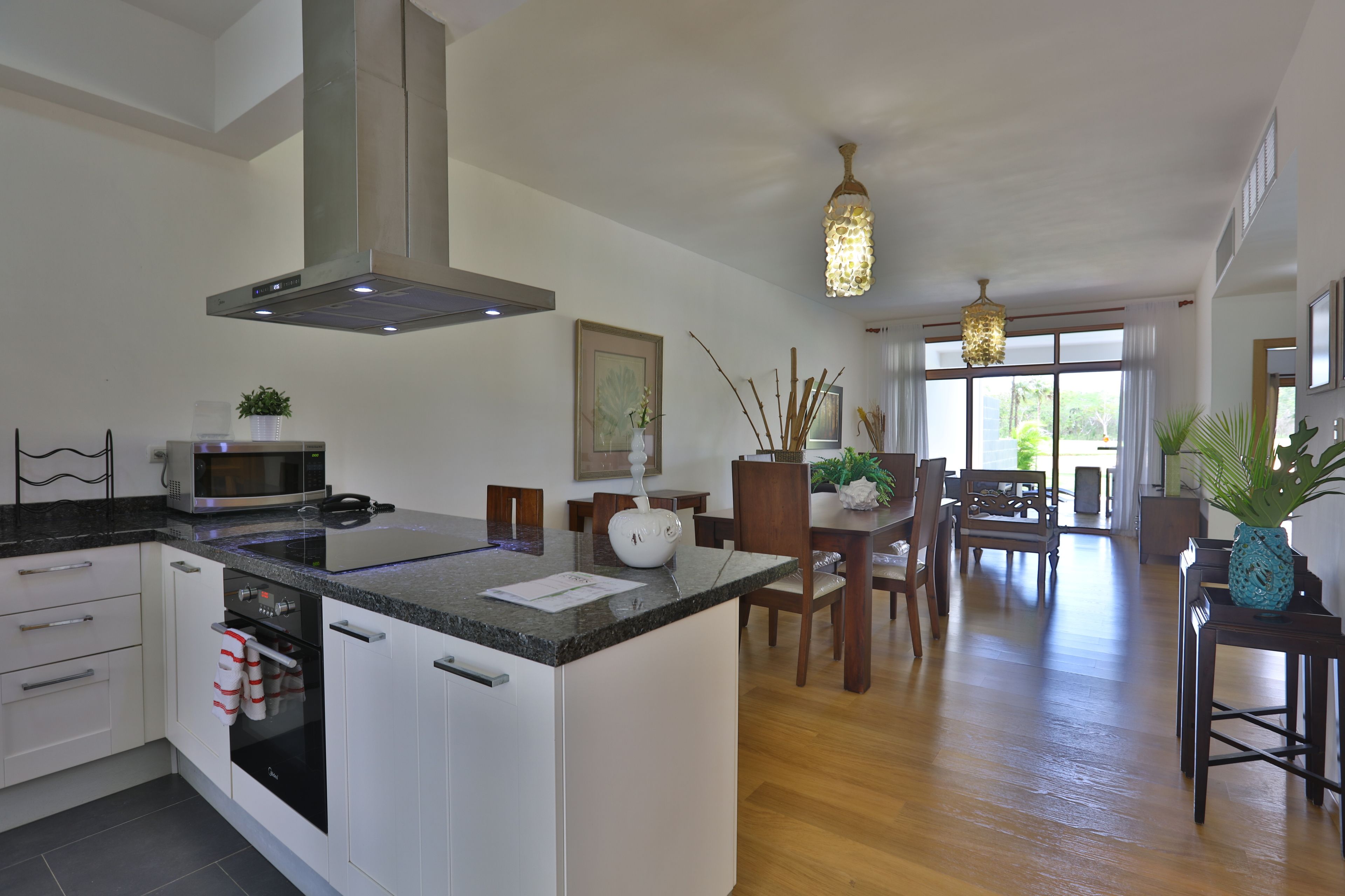 A modern kitchen with a central island, built-in oven, and microwave. There is a dining area with wooden furniture and a chandelier.