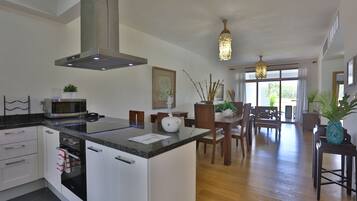 A modern kitchen with a central island, built-in oven, and microwave. There is a dining area with wooden furniture and a chandelier.