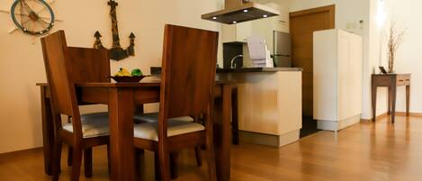 A dining area with a wooden table and chairs, a kitchen in the background, and a wooden floor.