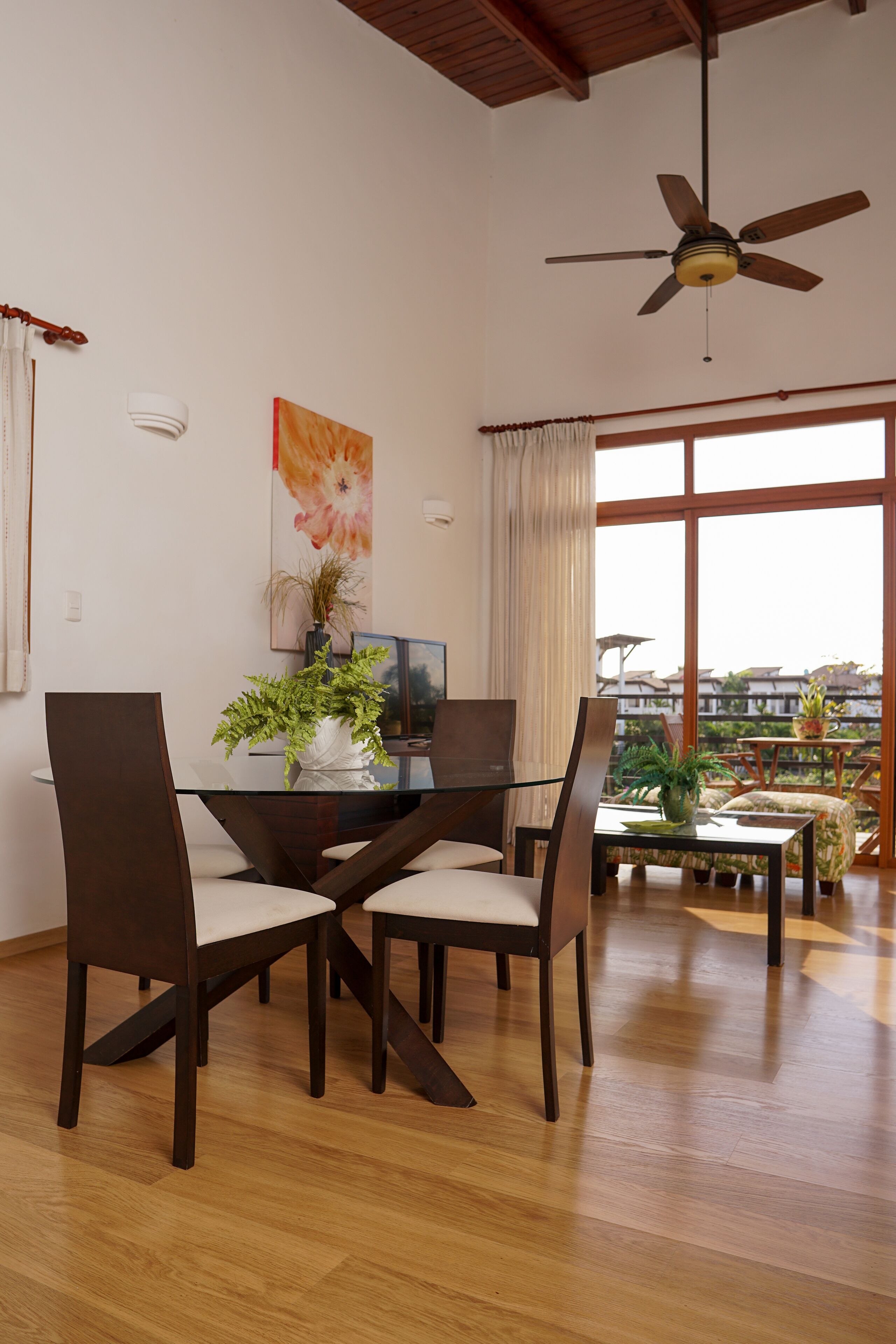 A dining area with a glass table, dark wooden chairs, and a ceiling fan.