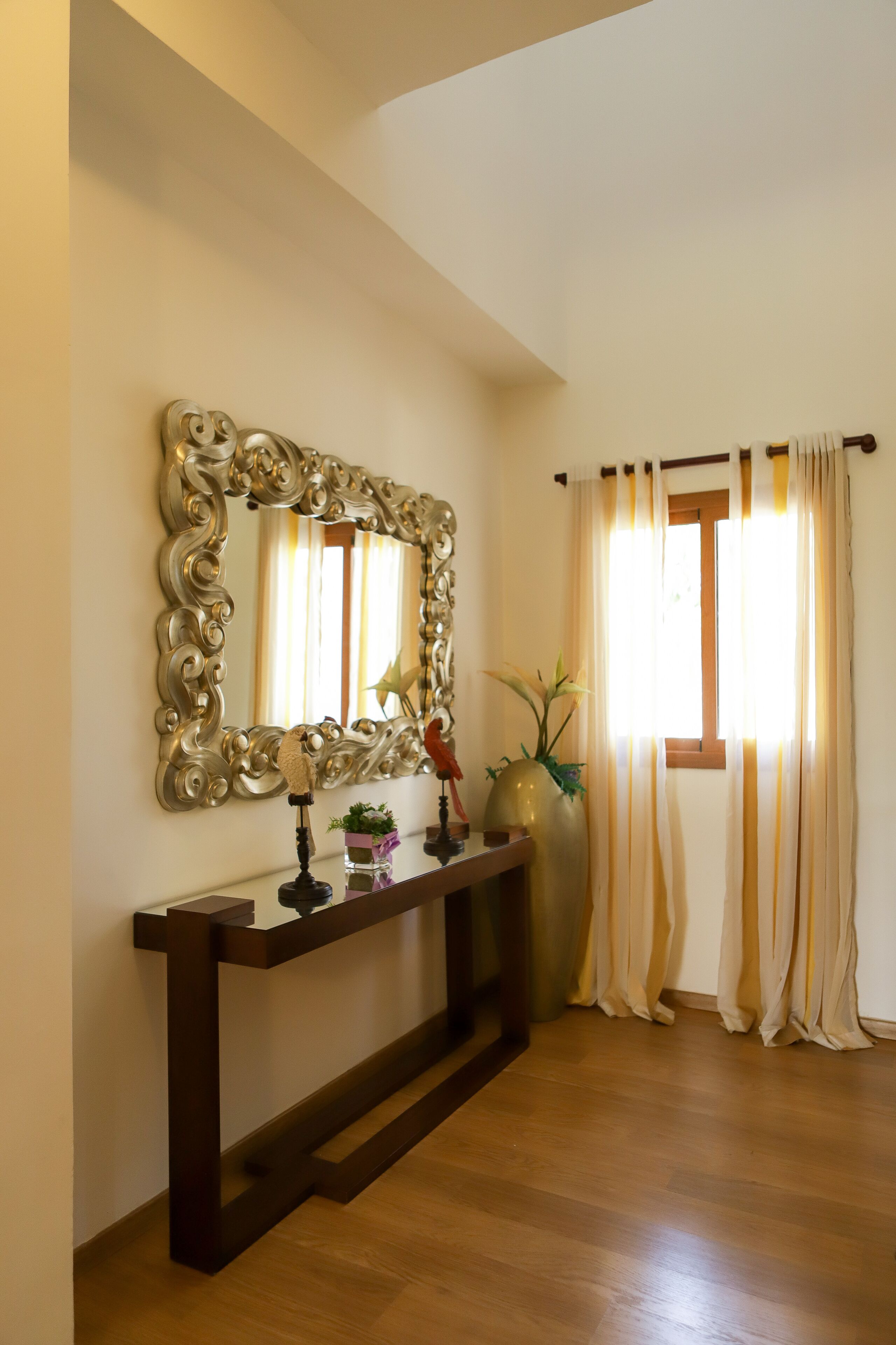 A room with a large ornate mirror, a console table with decorative items, a vase with plants, and a window with curtains.