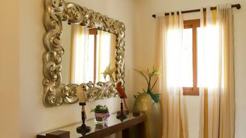 A room with a large ornate mirror, a console table with decorative items, a vase with plants, and a window with curtains.