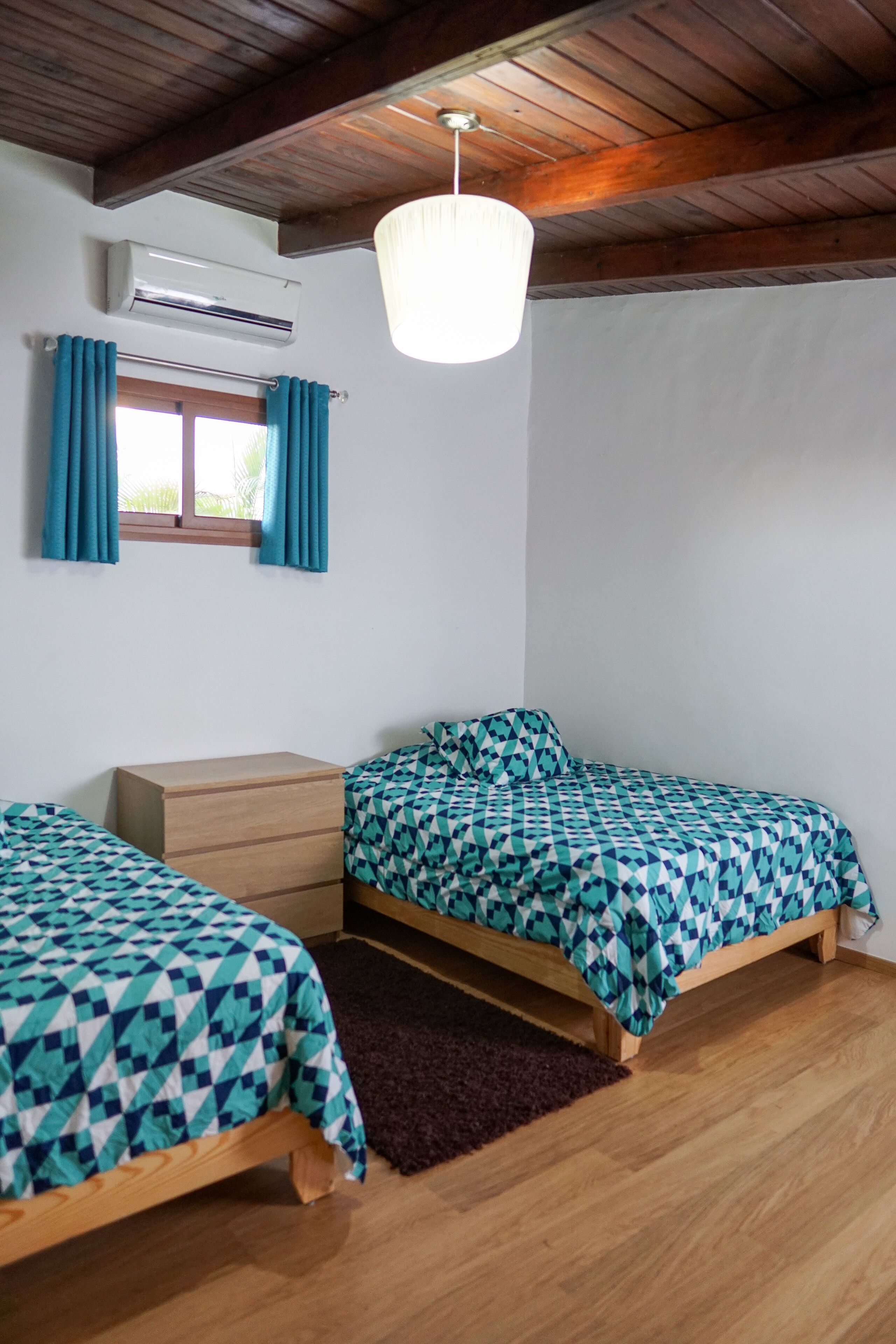 A bedroom with two single beds, a wooden dresser, an air conditioning unit, a window with curtains, and a hanging light fixture.