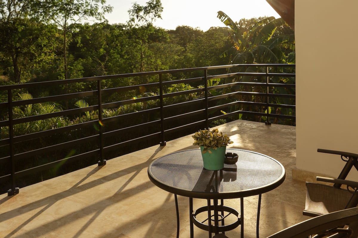 An outdoor patio with a round table and chairs, surrounded by a metal railing, overlooking a lush green landscape.