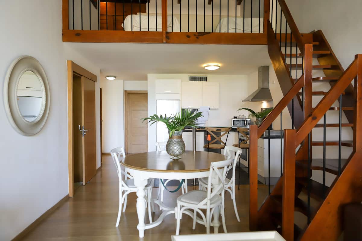 A dining area with a wooden table and white chairs, a wooden staircase, and a kitchen in the background.