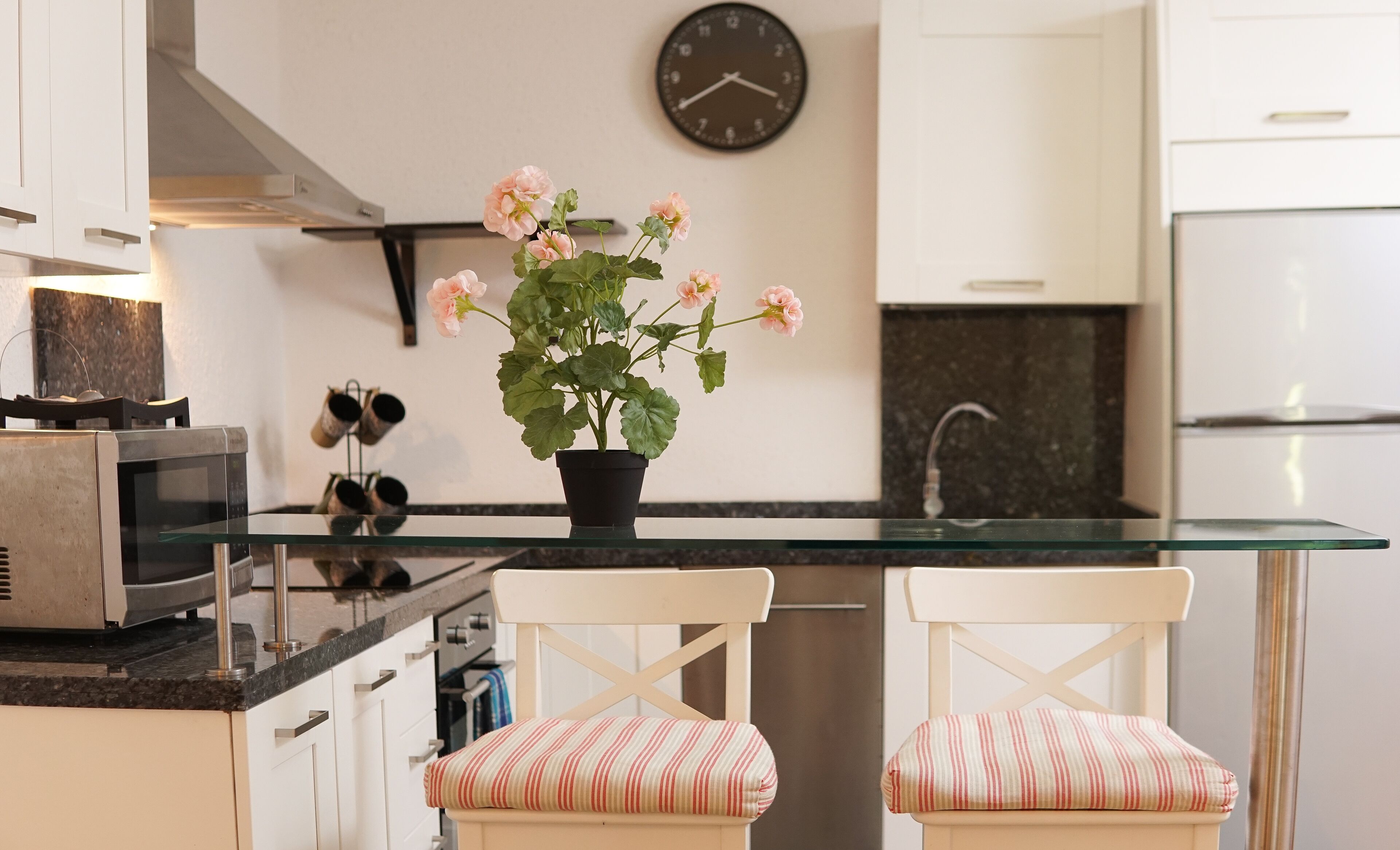 A modern kitchen with a glass island, two striped cushioned chairs, a potted plant with flowers, a clock, and stainless steel appliances.