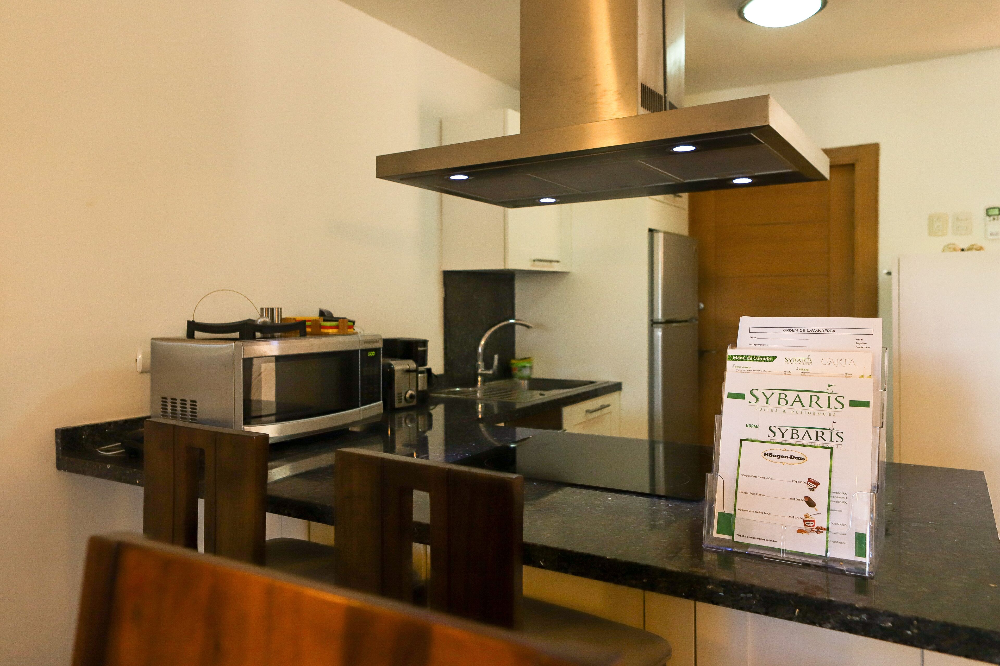 Een moderne keuken met een roestvrijstalen afzuigkap, een magnetron, een broodrooster en een koelkast. Op de voorgrond staan stoelen en een tafel, en een deur die leidt naar een andere kamer.