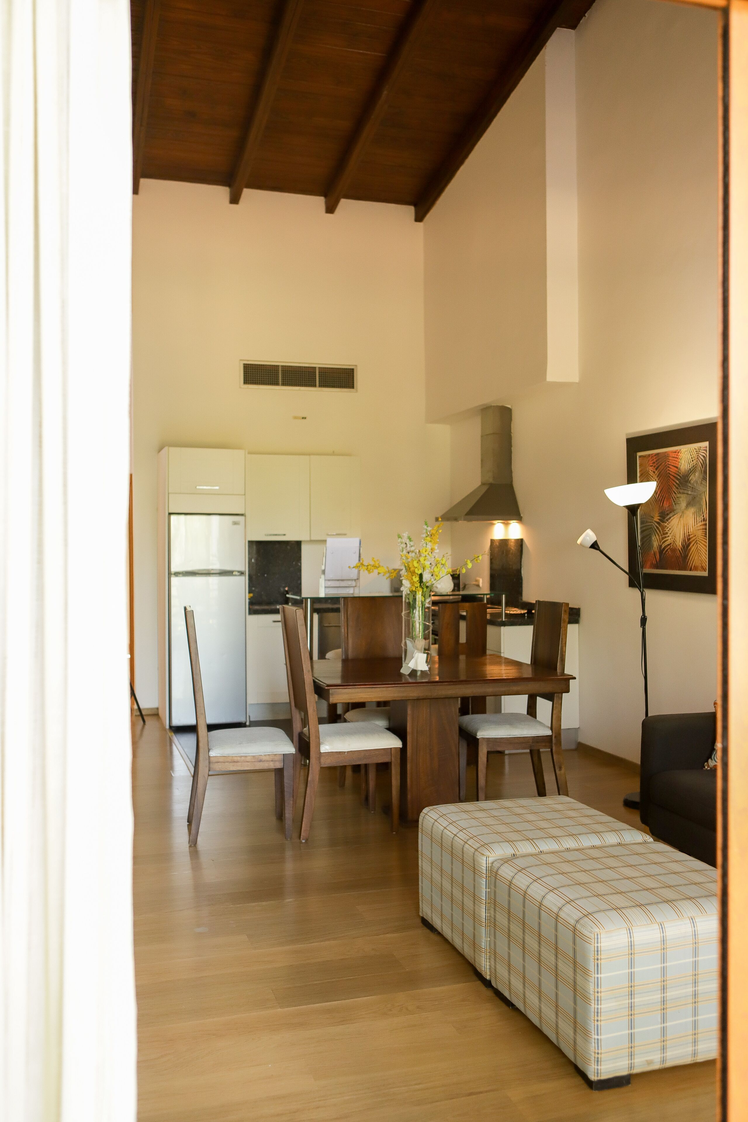 Une cuisine compacte avec des meubles blancs, un réfrigérateur et un four à micro-ondes. On y trouve une table à manger avec des chaises et un banc. Une lampe sur pied et un tableau encadré sont accrochés au mur.