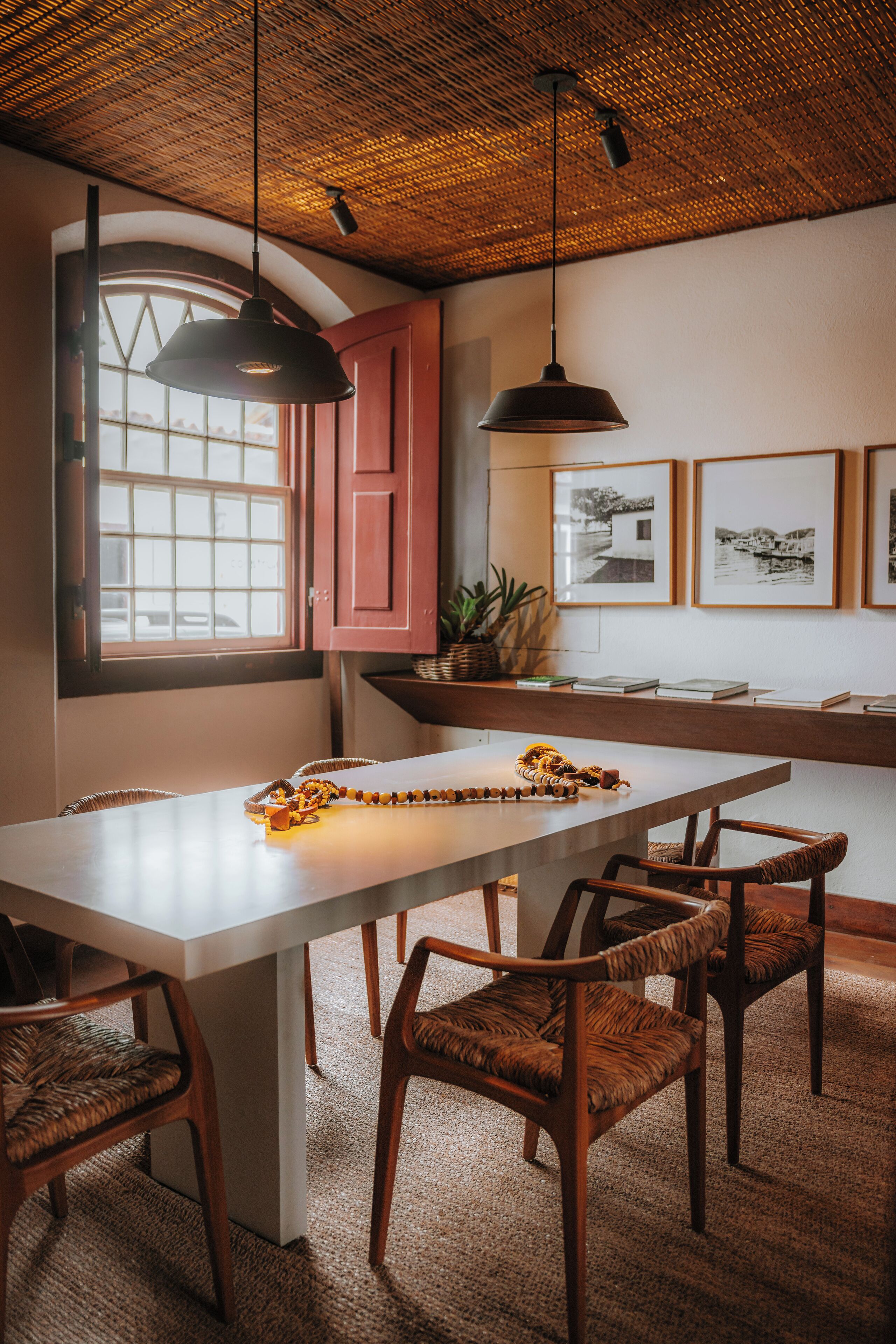 Un comedor con una mesa grande, sillas de mimbre, lámparas colgantes, una ventana con vistas y cuadros en la pared.