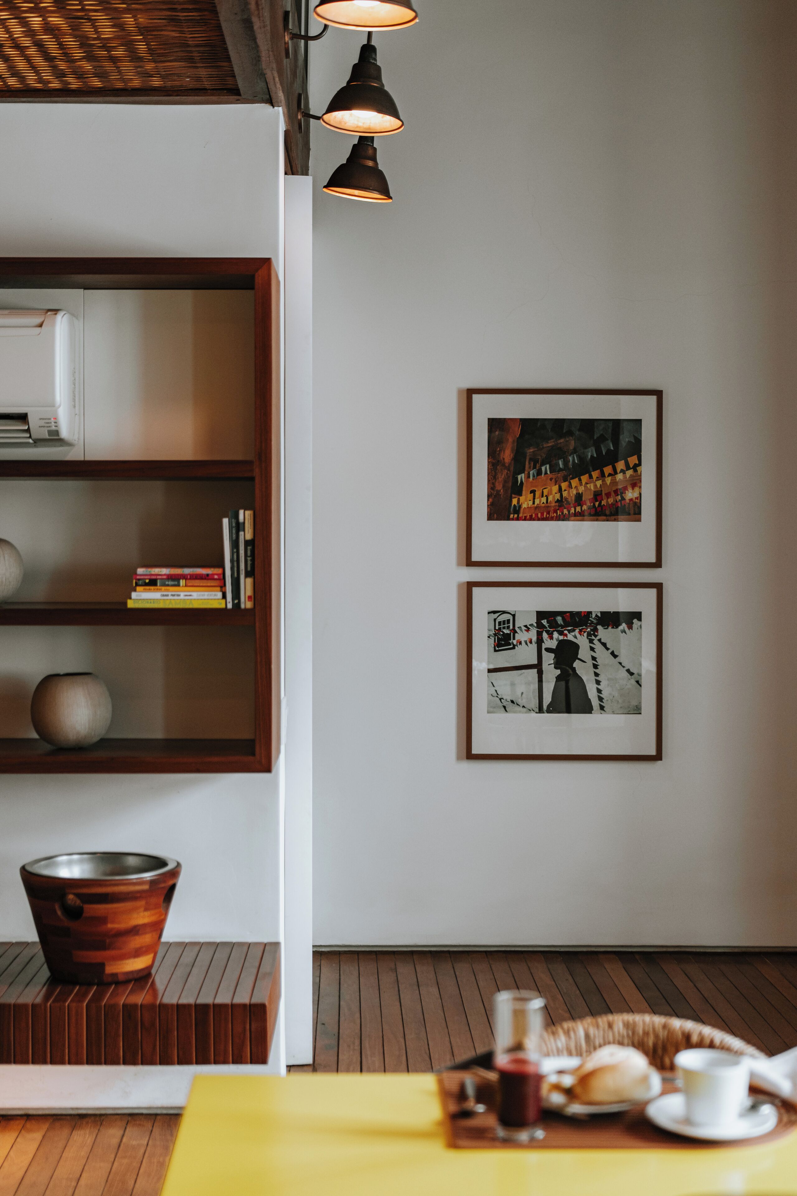 A room with a wooden floor, a yellow table with breakfast items, a bookshelf with books and a vase, and two framed pictures on the wall.