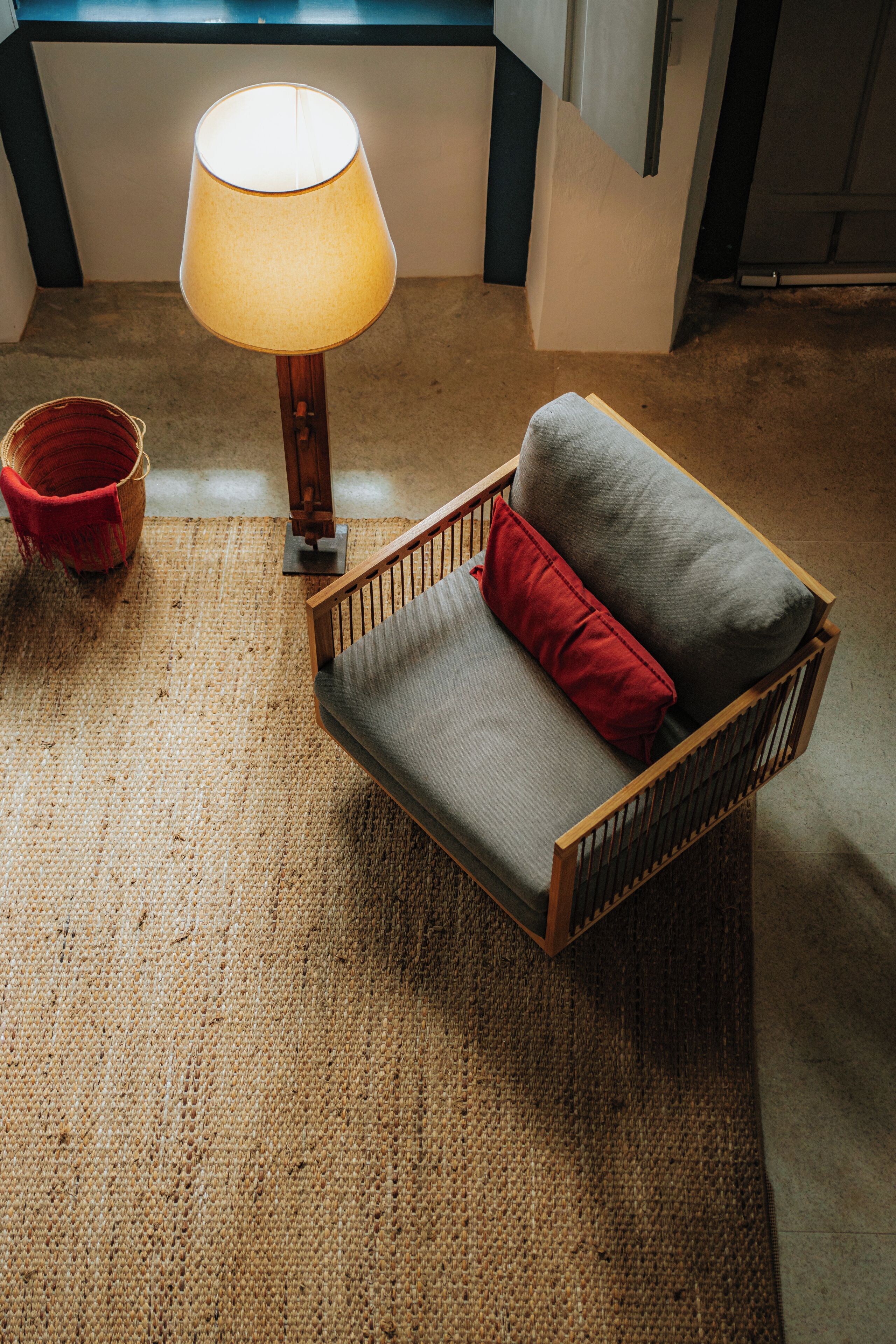 Una habitación con un sofá de madera, un cojín rojo, una lámpara y una alfombra tejida.