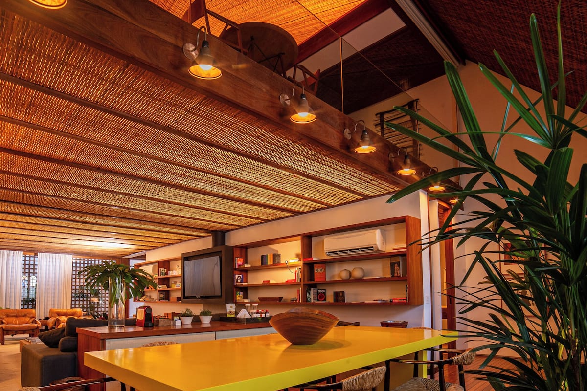 A modern living room with a wooden floor, a yellow dining table, and several chairs. There is a television mounted on the wall, and a large potted plant is visible.