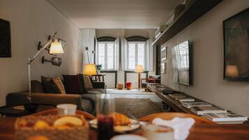 Una sala de estar acogedora con un sofá, una mesita de centro con comida y bebidas, una estantería y vistas a través de una puerta.
