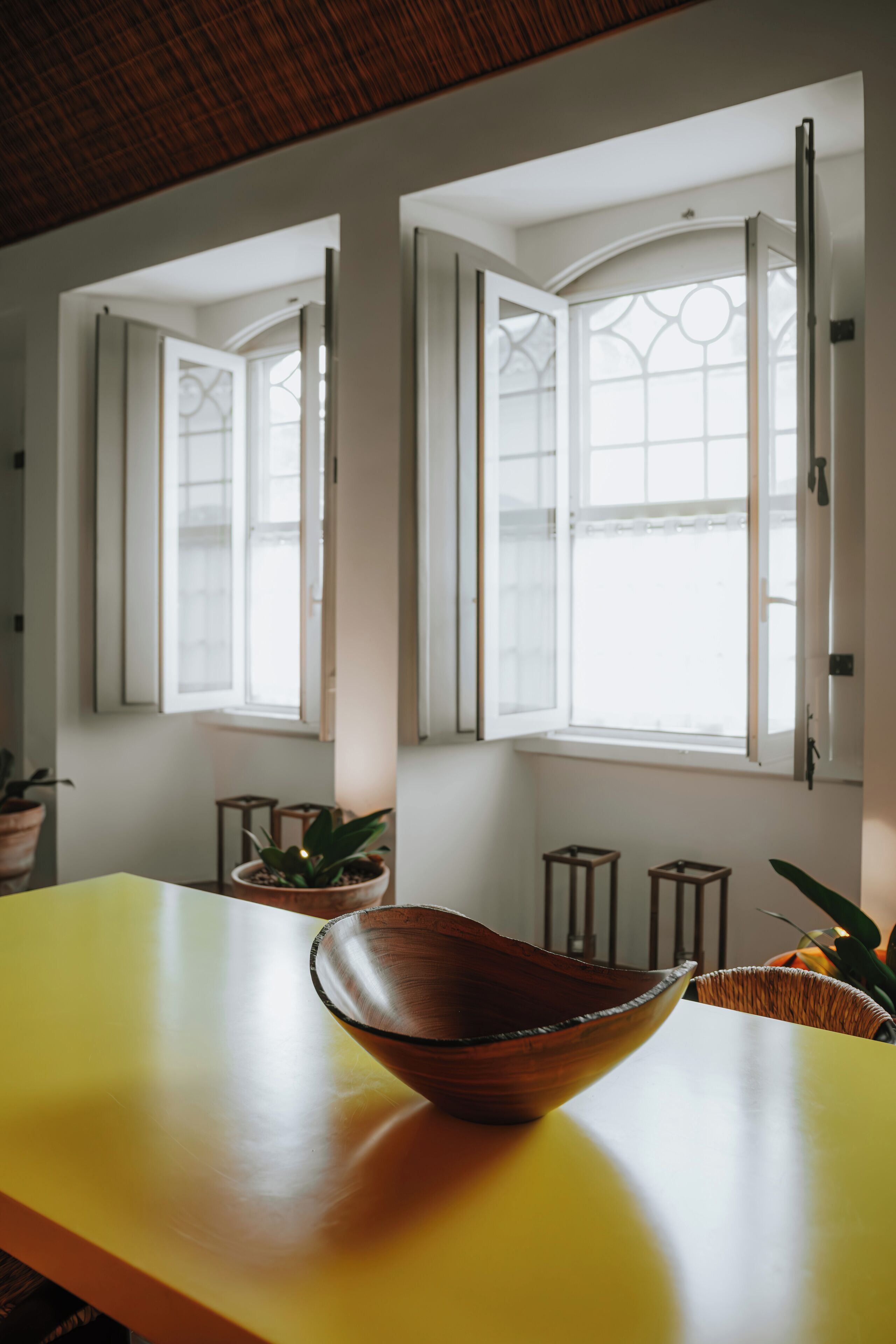 Um quarto com uma mesa amarela, uma tigela de madeira e duas janelas abertas com molduras brancas e painéis de vidro decorativos.