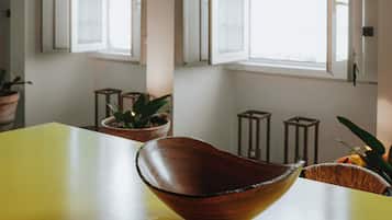Um quarto com uma mesa amarela, uma tigela de madeira e duas janelas abertas com molduras brancas e painéis de vidro decorativos.