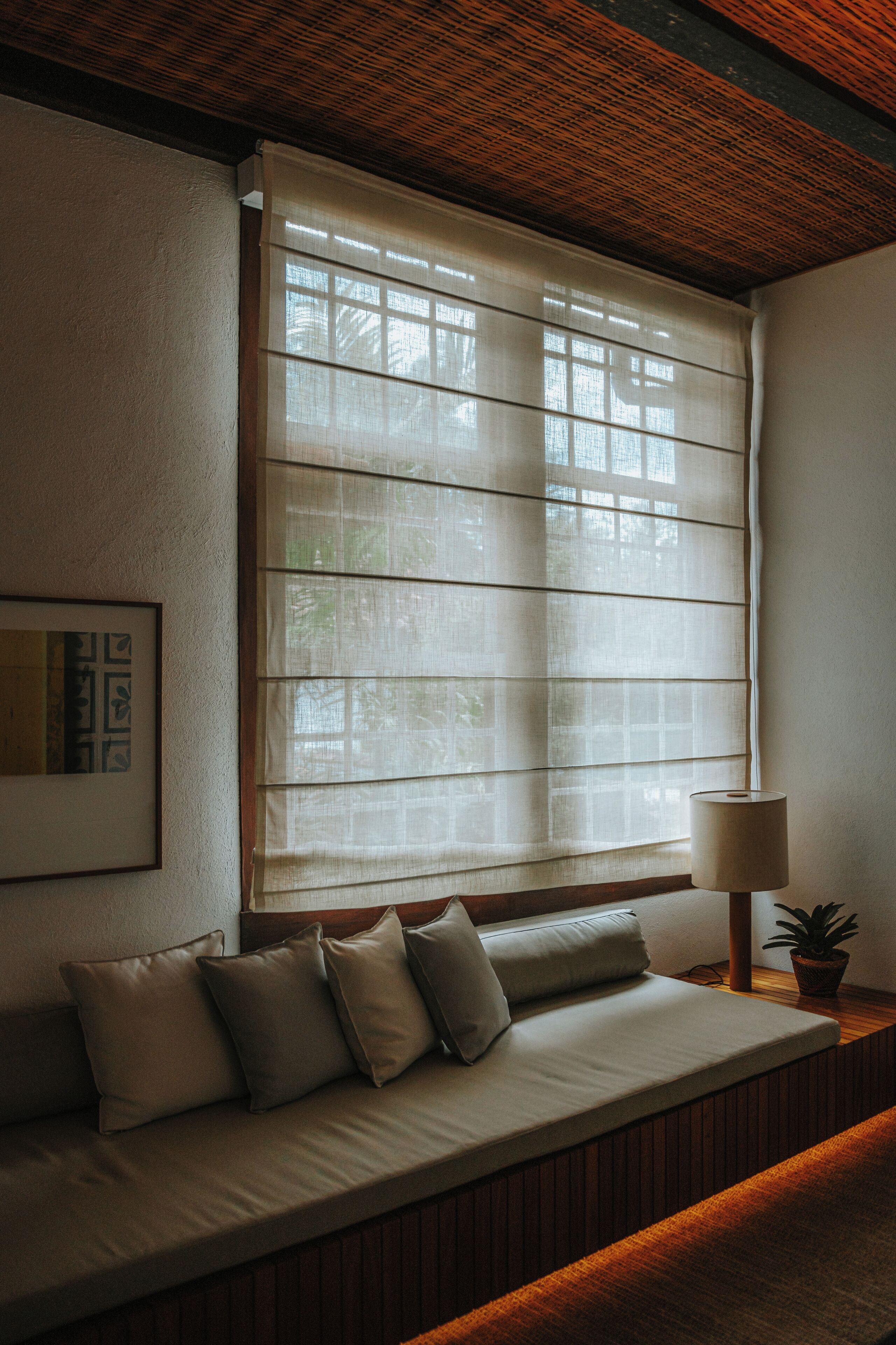A room with a window covered by blinds, a sofa with cushions, a lamp, and a framed picture on the wall.