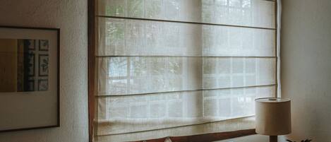 A room with a window covered by blinds, a sofa with cushions, a lamp, and a framed picture on the wall.