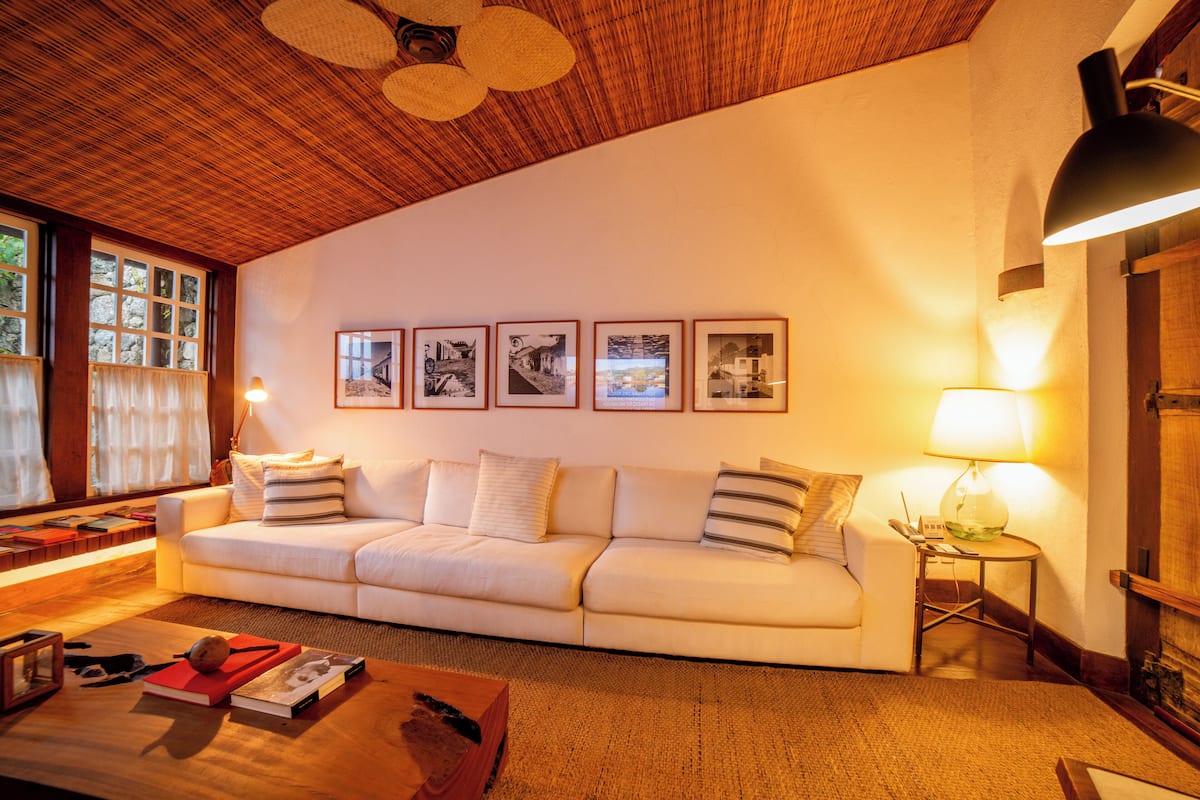 A cozy living room with a wooden ceiling, a white sofa, framed pictures on the wall, and a lamp on a side table.