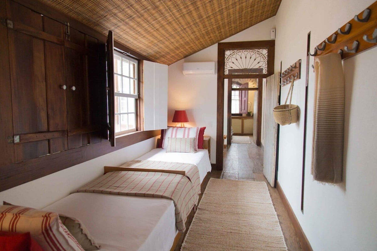 A bedroom with three beds, a wooden ceiling, a window, a lamp, and a hallway leading to a kitchen.