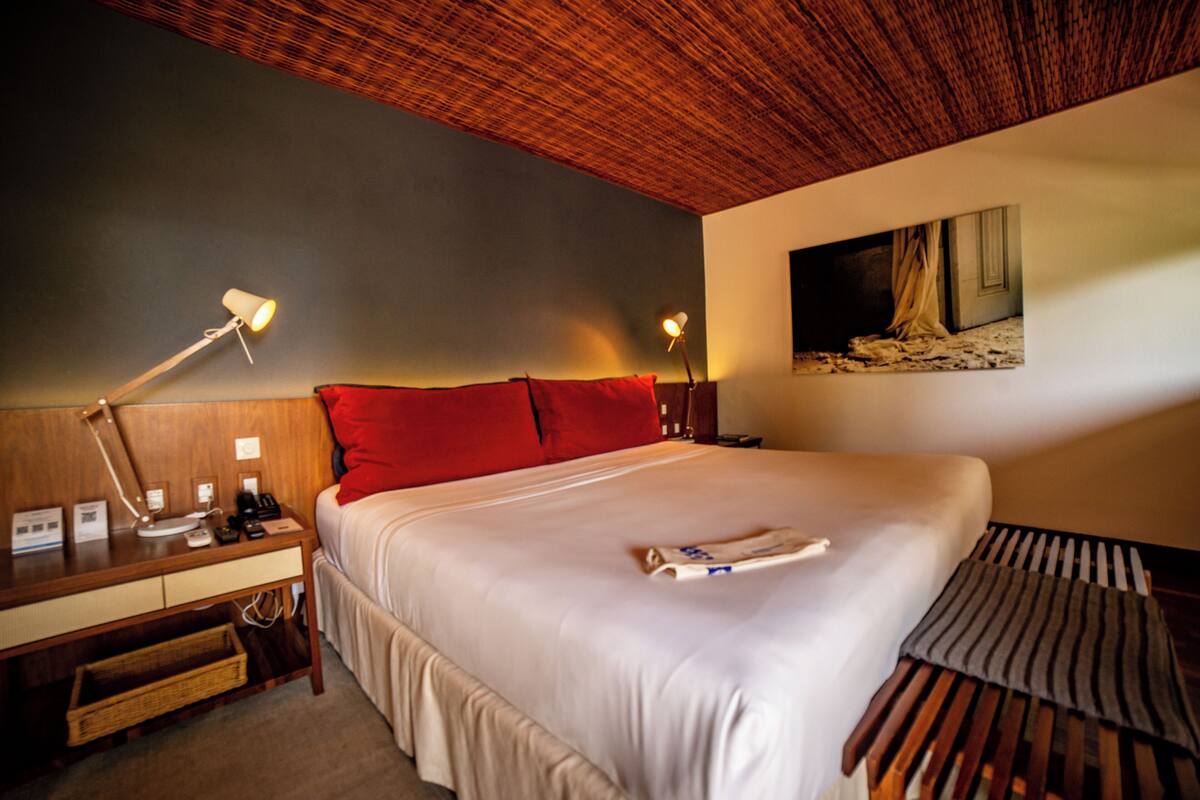 A hotel room with a large bed, a desk with a lamp, a bedside table with a phone, and a painting on the wall.