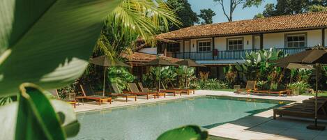 A pool area with lounge chairs and umbrellas, surrounded by tropical plants and a building with a tiled roof.