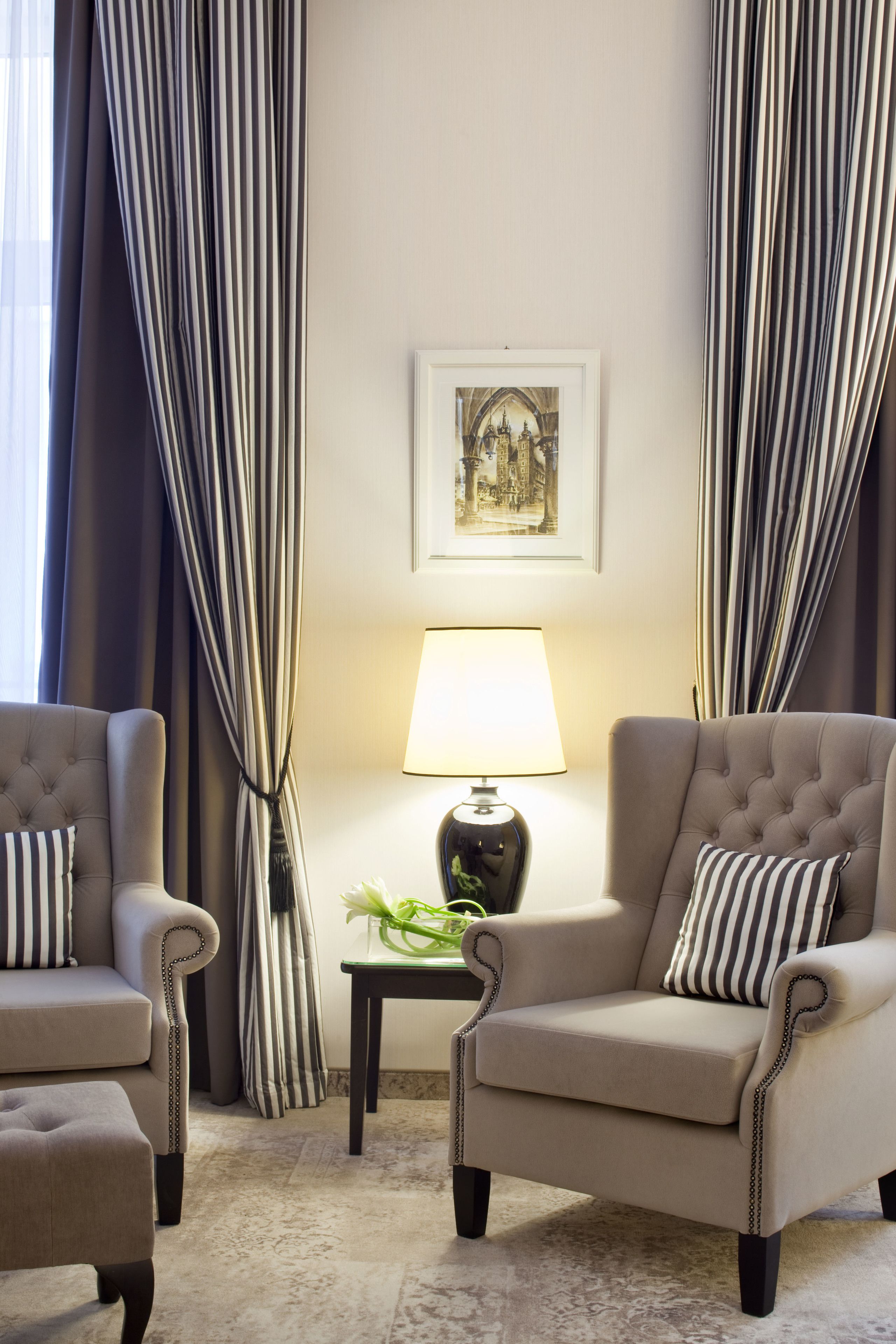 A room with two armchairs, a small table with a lamp, a framed picture on the wall, and striped curtains.