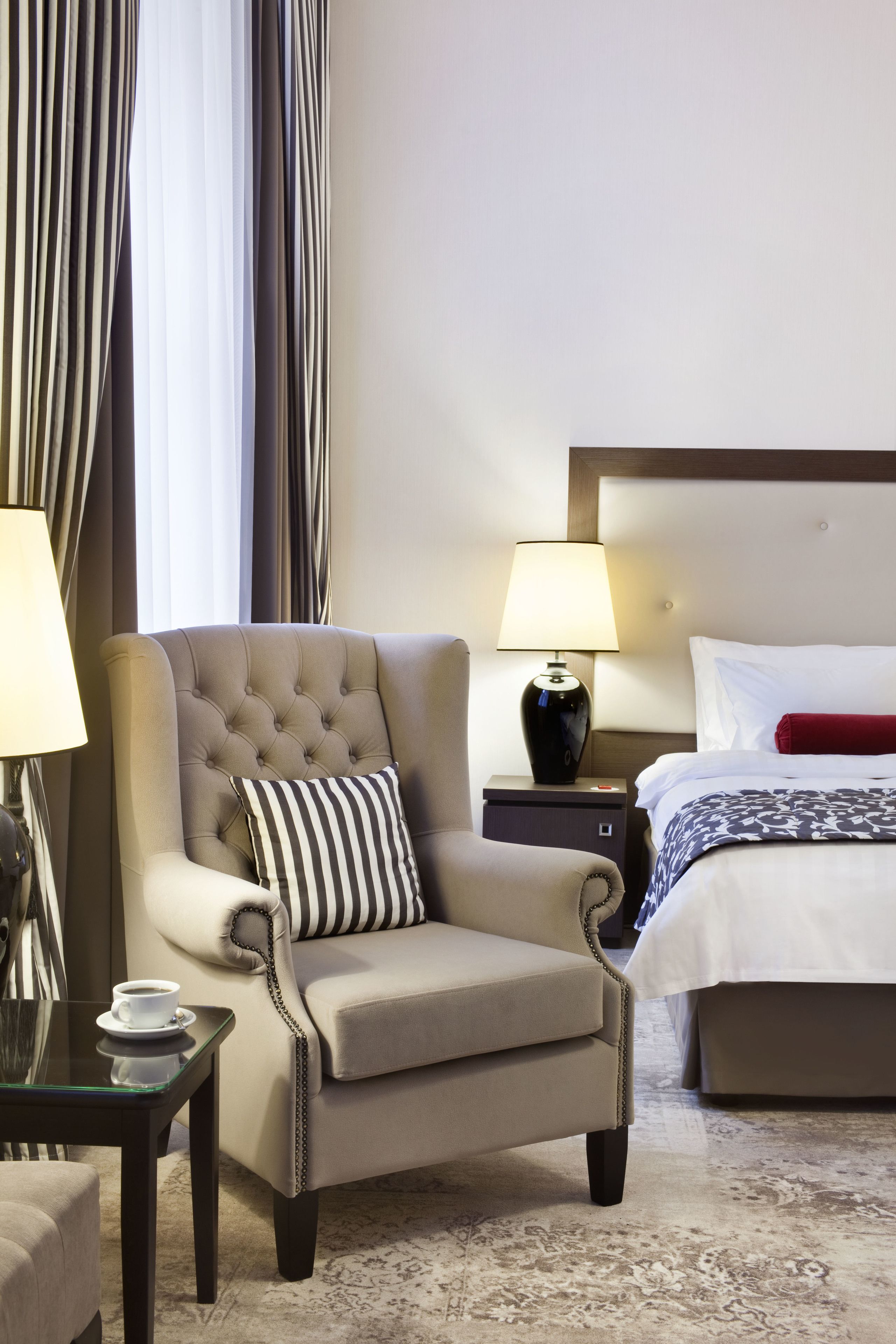 A hotel room with a tufted armchair, a bedside table with a lamp, a bed with white bedding, and a patterned rug.