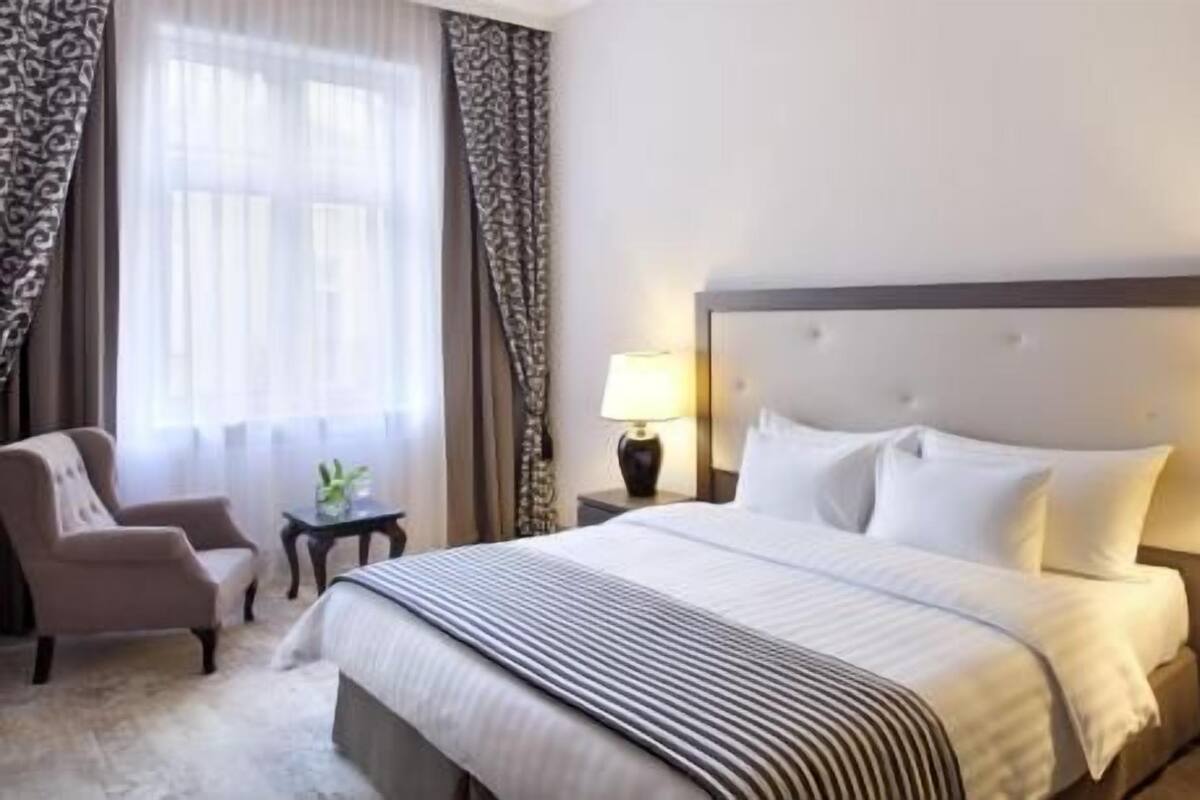 Ein Hotelzimmer mit Bett, Sessel, Nachttisch, Lampe und einem Fenster mit Vorhängen.