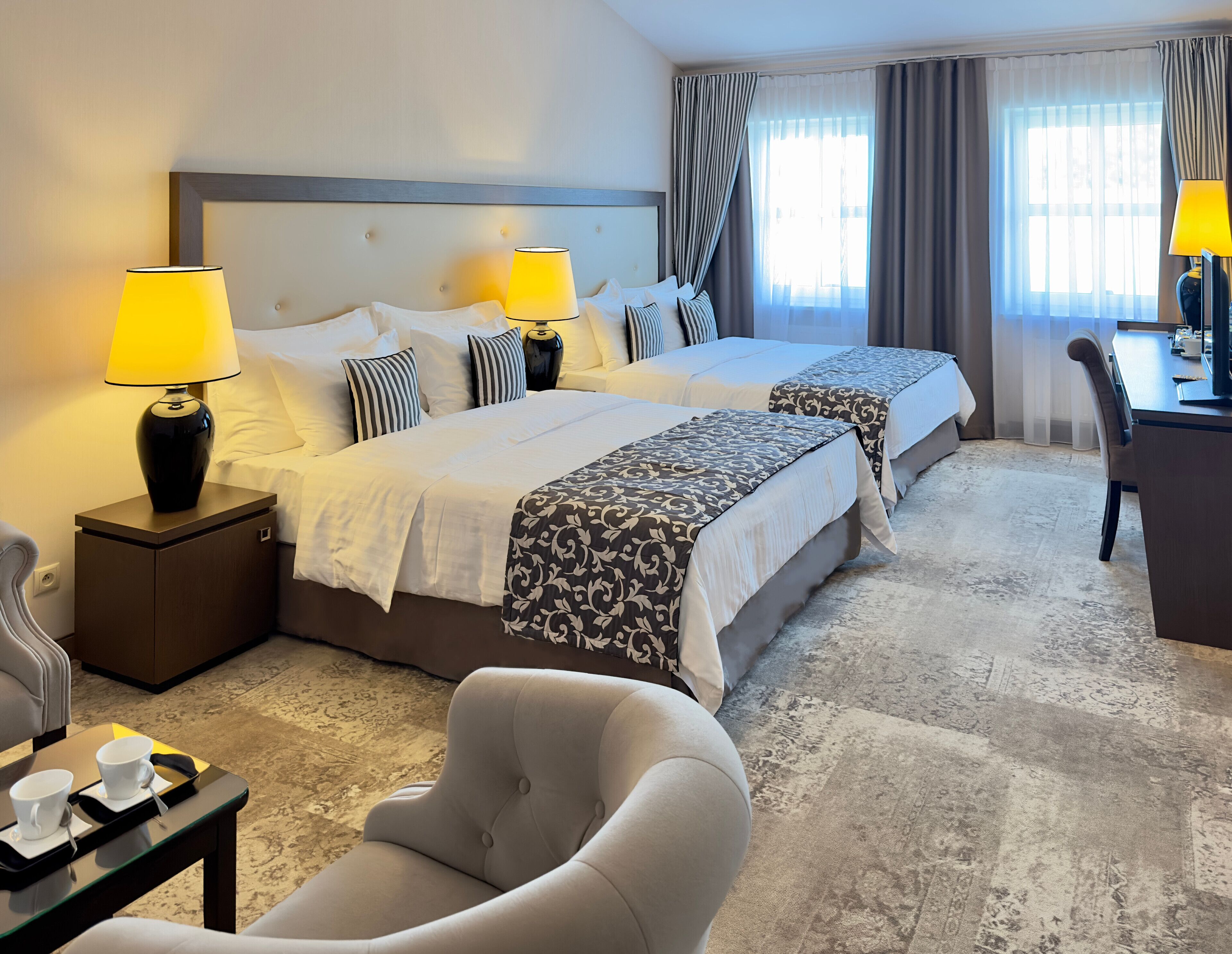 A hotel room with two beds, a desk, a chair, and a lamp.