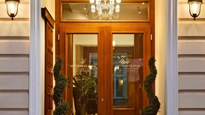Entrada de hotel con puerta de madera, paneles de vidrio y una araña. Hay dos plantas en macetas en la entrada.