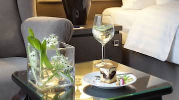 A hotel room with a bed, a nightstand with a lamp, a glass table with a glass of water and a plate with food, and a chair.