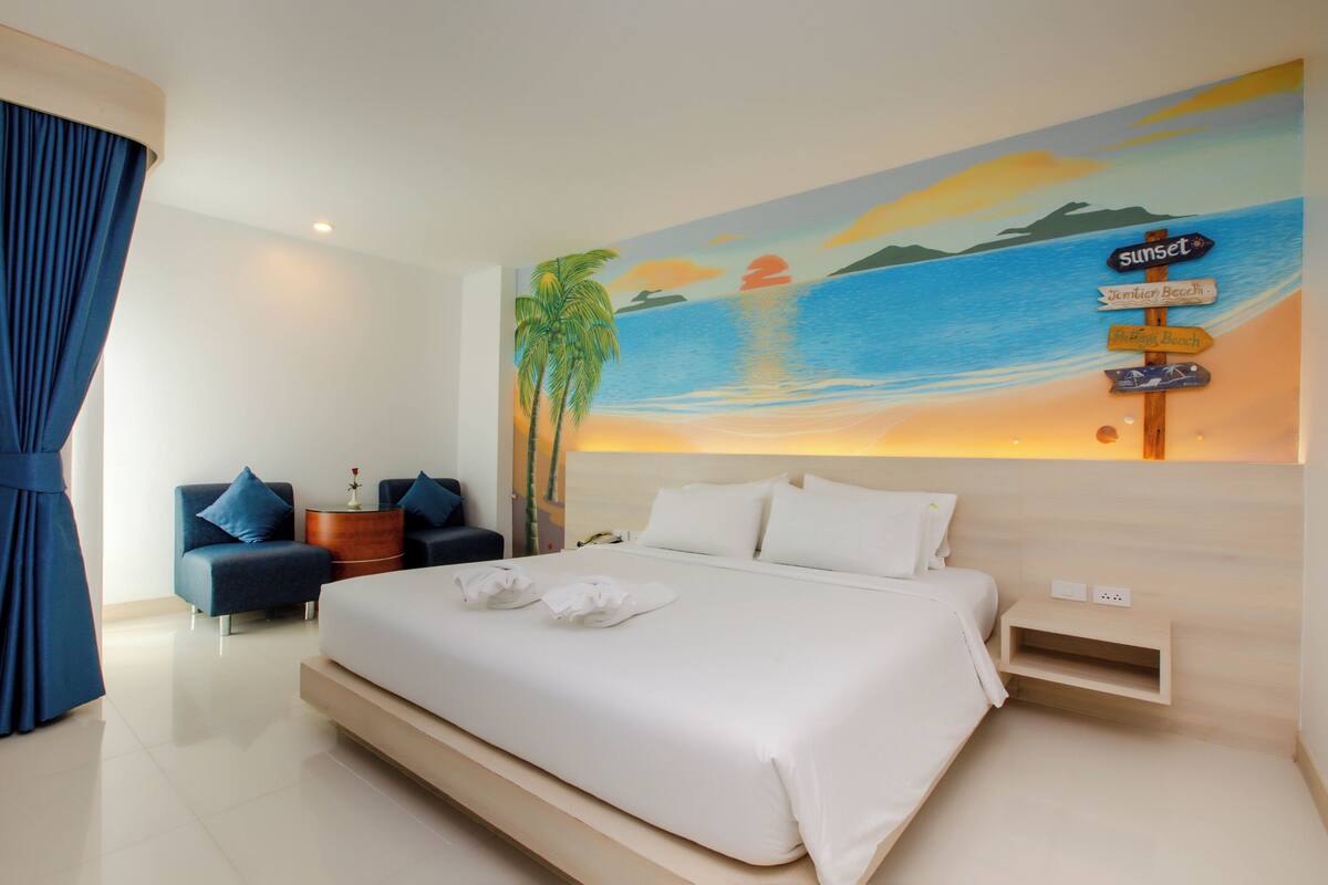 A modern hotel room with a large bed, a nightstand, a desk, and a wall mural depicting a sunset over the ocean.