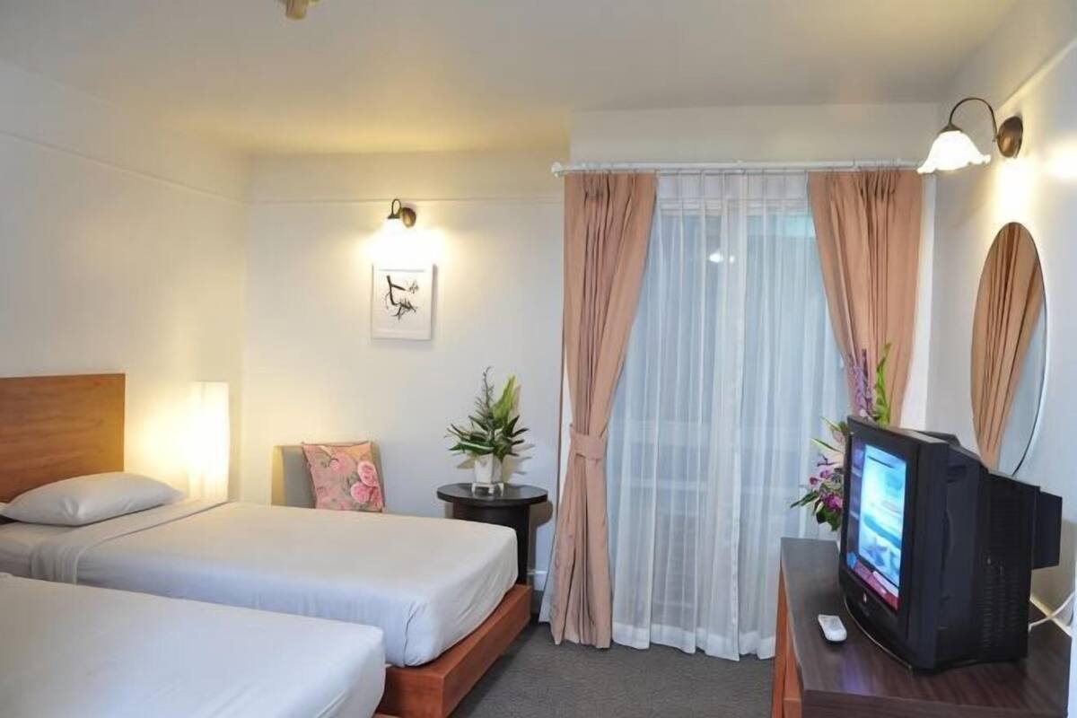 A hotel room with two beds, a TV, a small table with a plant, and a window with sheer curtains.