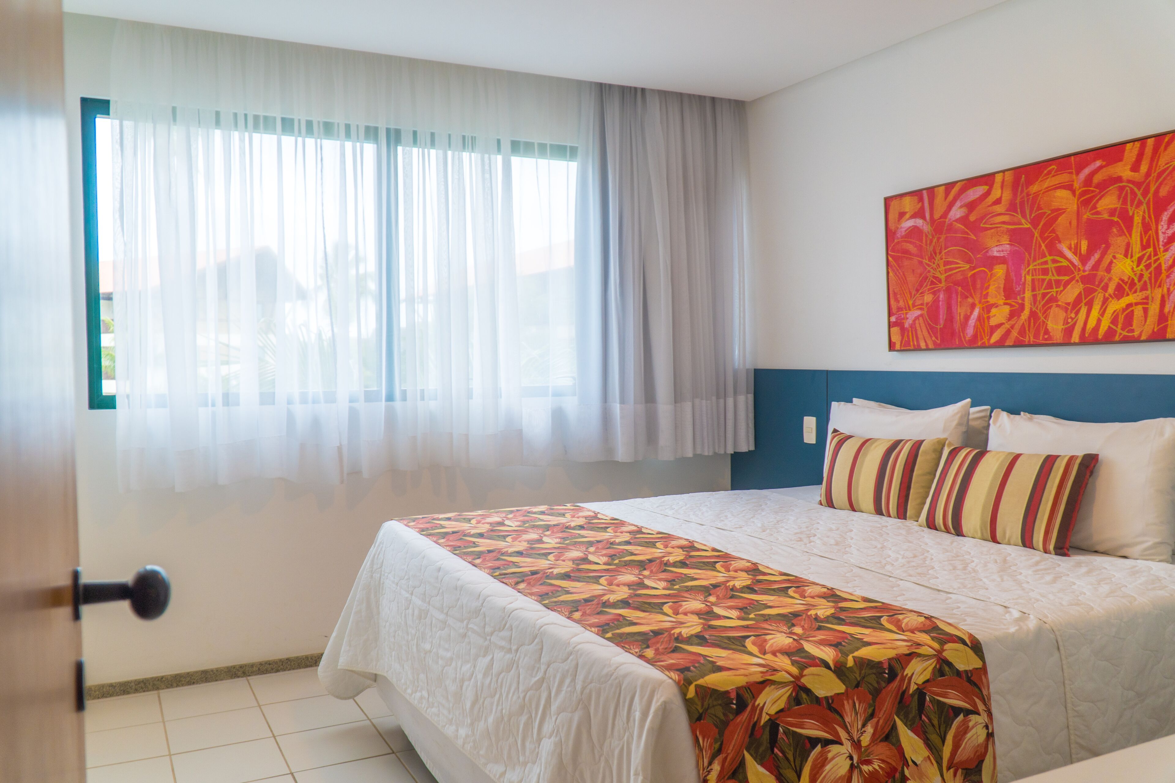 Habitación de hotel con cama, ventana con cortinas traslúcidas, un cuadro abstracto colorido en la pared y una puerta con pomo redondo.