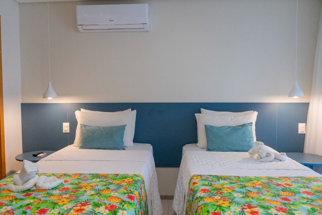 Een hotelkamer met twee bedden, elk voorzien van een bloemenprint deken, witte kussens en een blauw hoofdeinde. Daarboven hangt een airconditioning en aan de linkerkant staat een klein tafeltje met een afstandsbediening.