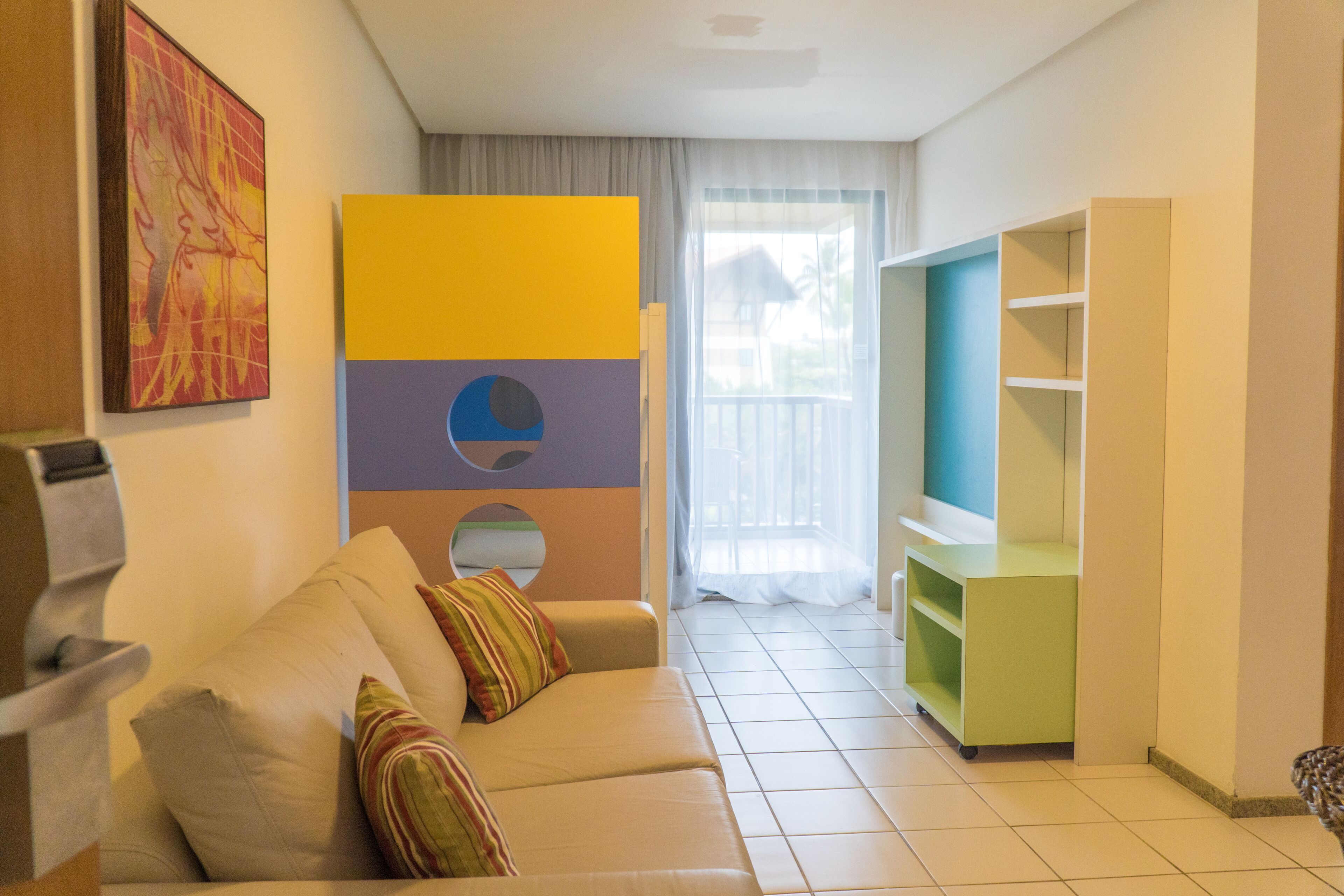A living room with a beige sofa, colorful cushions, a green entertainment unit, and a large window with a view.