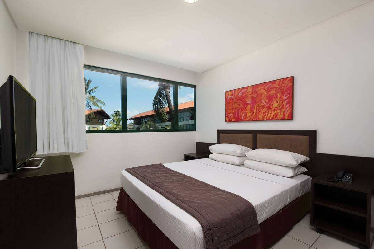 Ein Hotelzimmer mit Bett, Fernseher, Nachttisch, Telefon, Fenster mit Blick auf Palmen und Gebäude sowie einem roten, abstrakten Gemälde an der Wand.