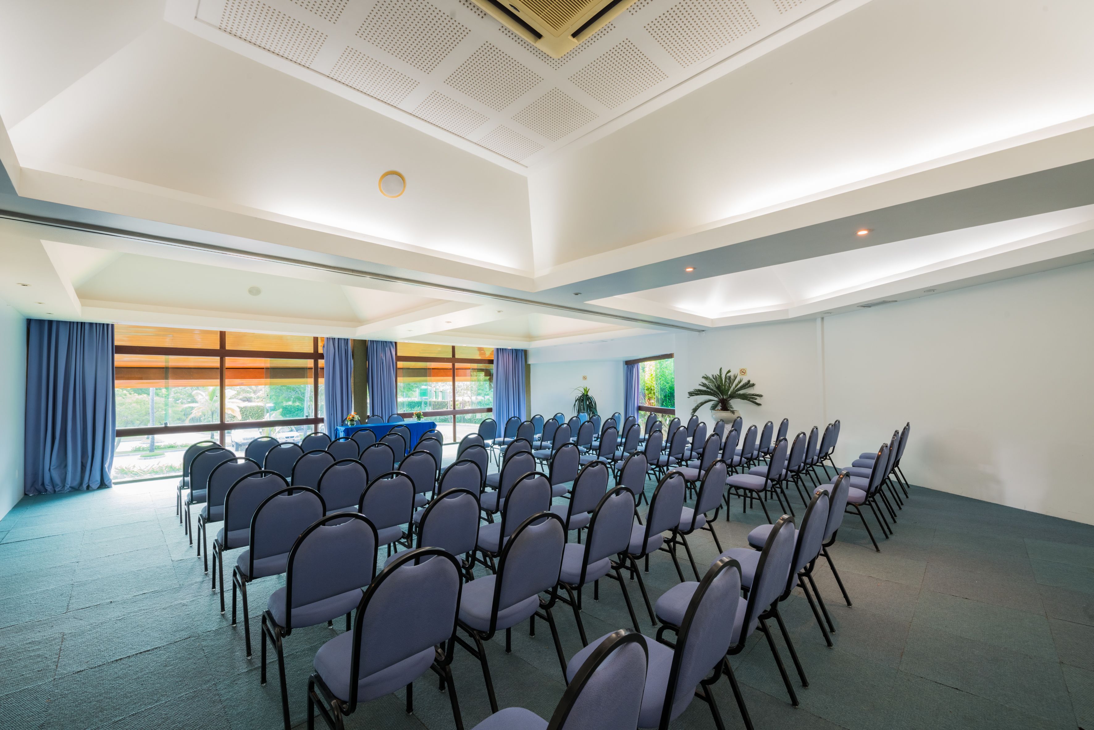 A conference room with rows of chairs, a blue table, and large windows with curtains.