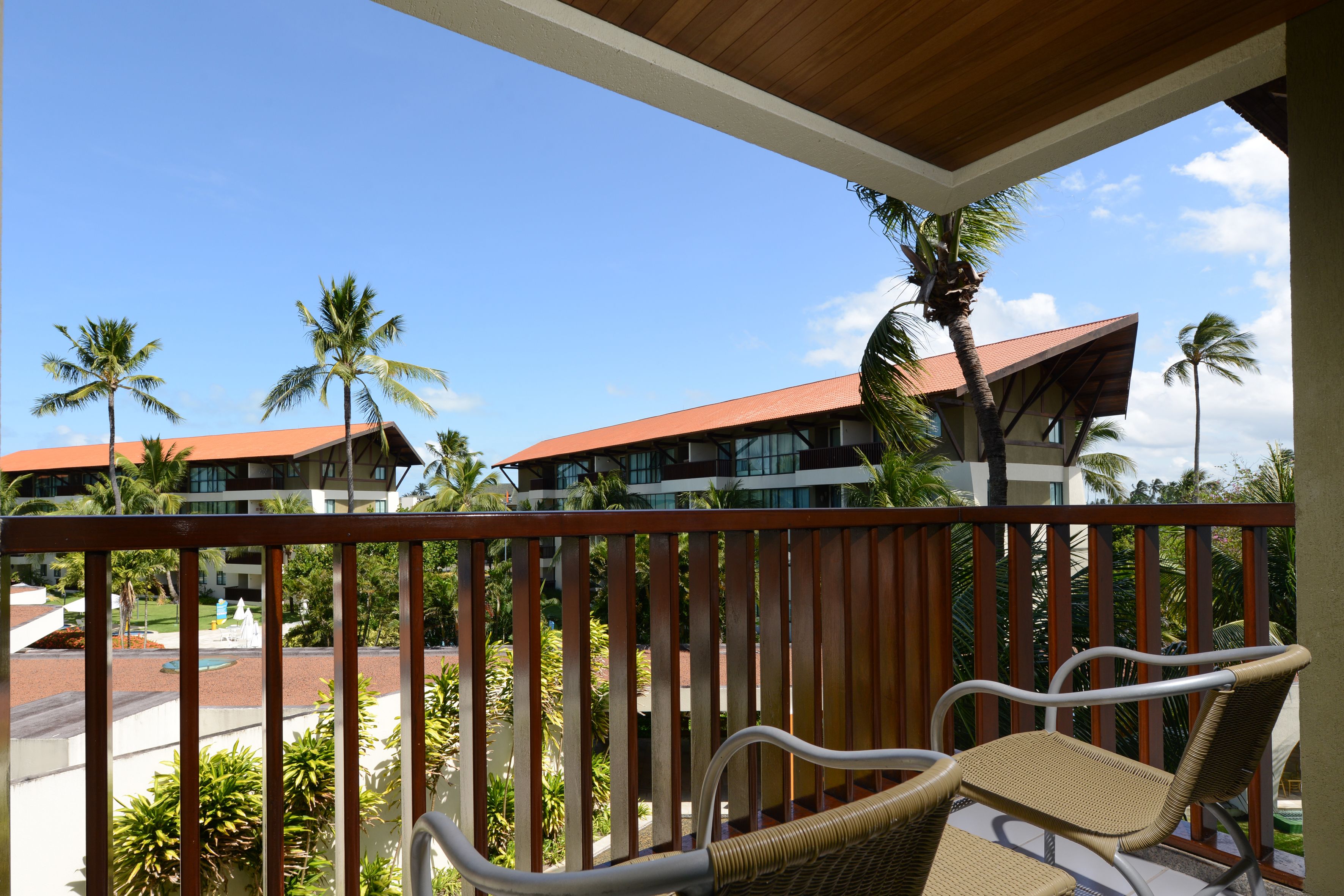 A balcony with wicker furniture, overlooking a pool and palm trees, with buildings in the background.