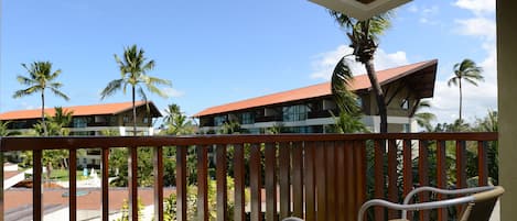 A balcony with wicker furniture, overlooking a pool and palm trees, with buildings in the background.