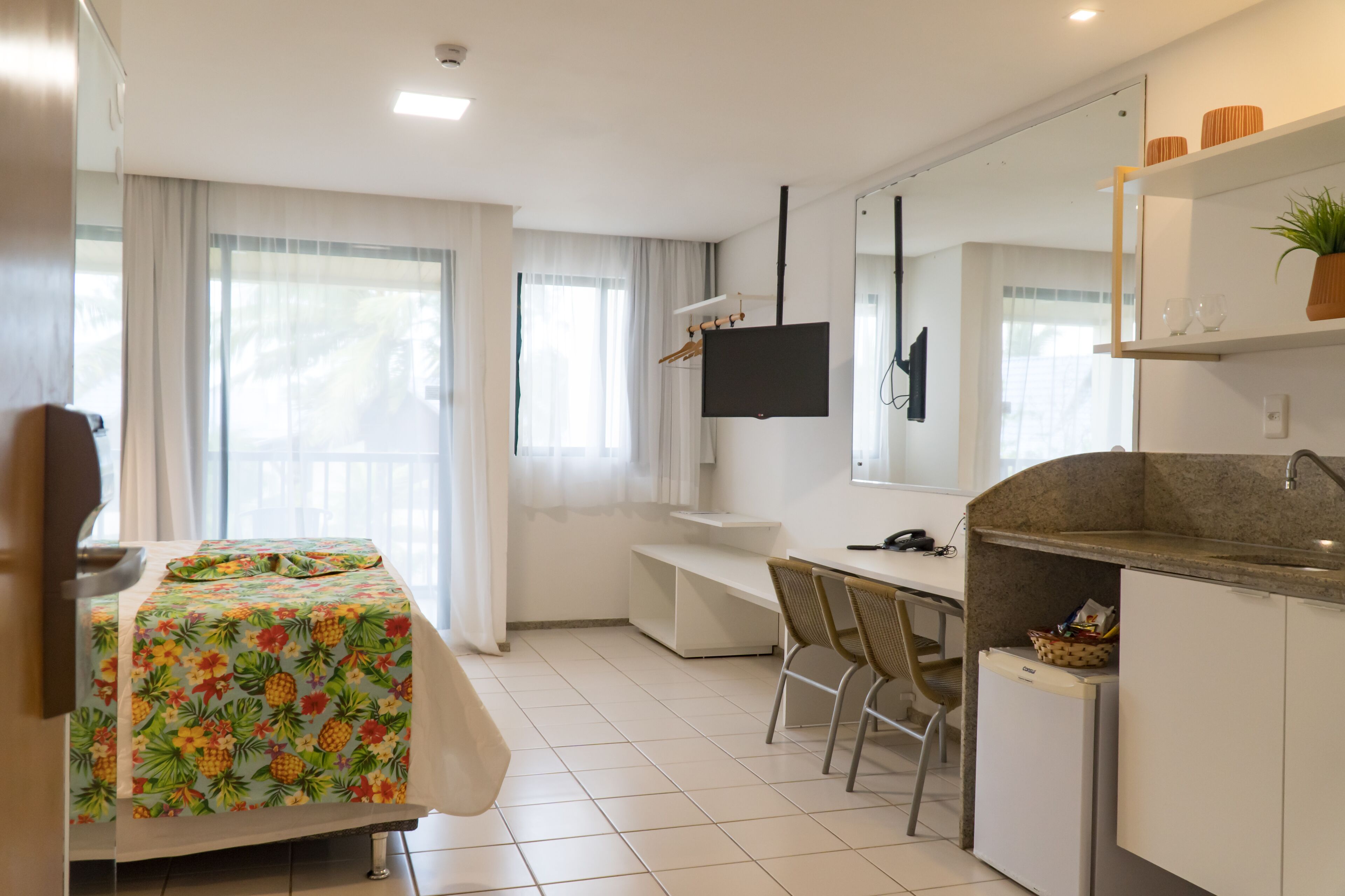 A hotel room with a bed, a desk, a chair, a TV, and a kitchenette.