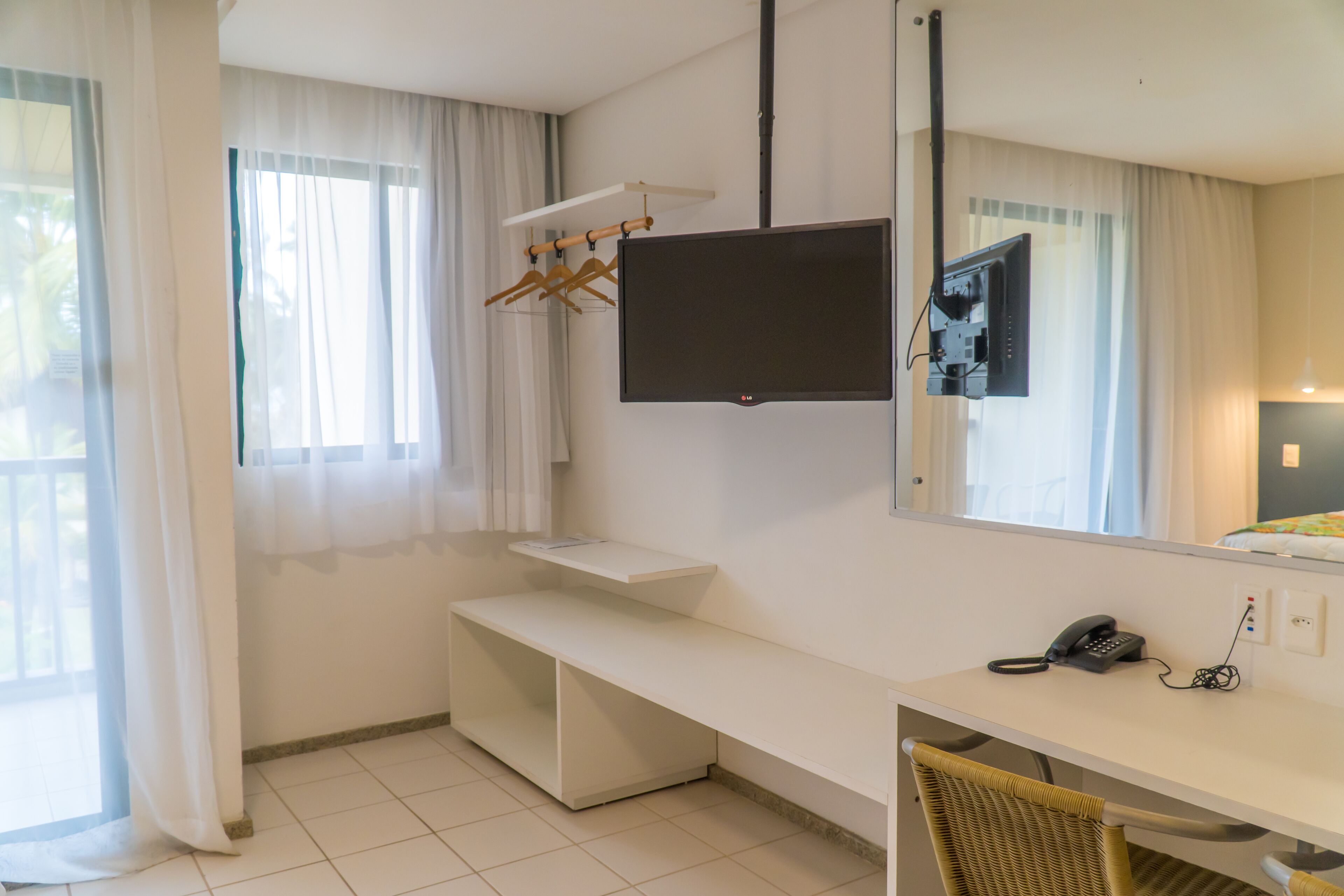 A hotel room with a white desk, a bench, a TV mounted on the wall, a telephone, and a hanging clothes rack.