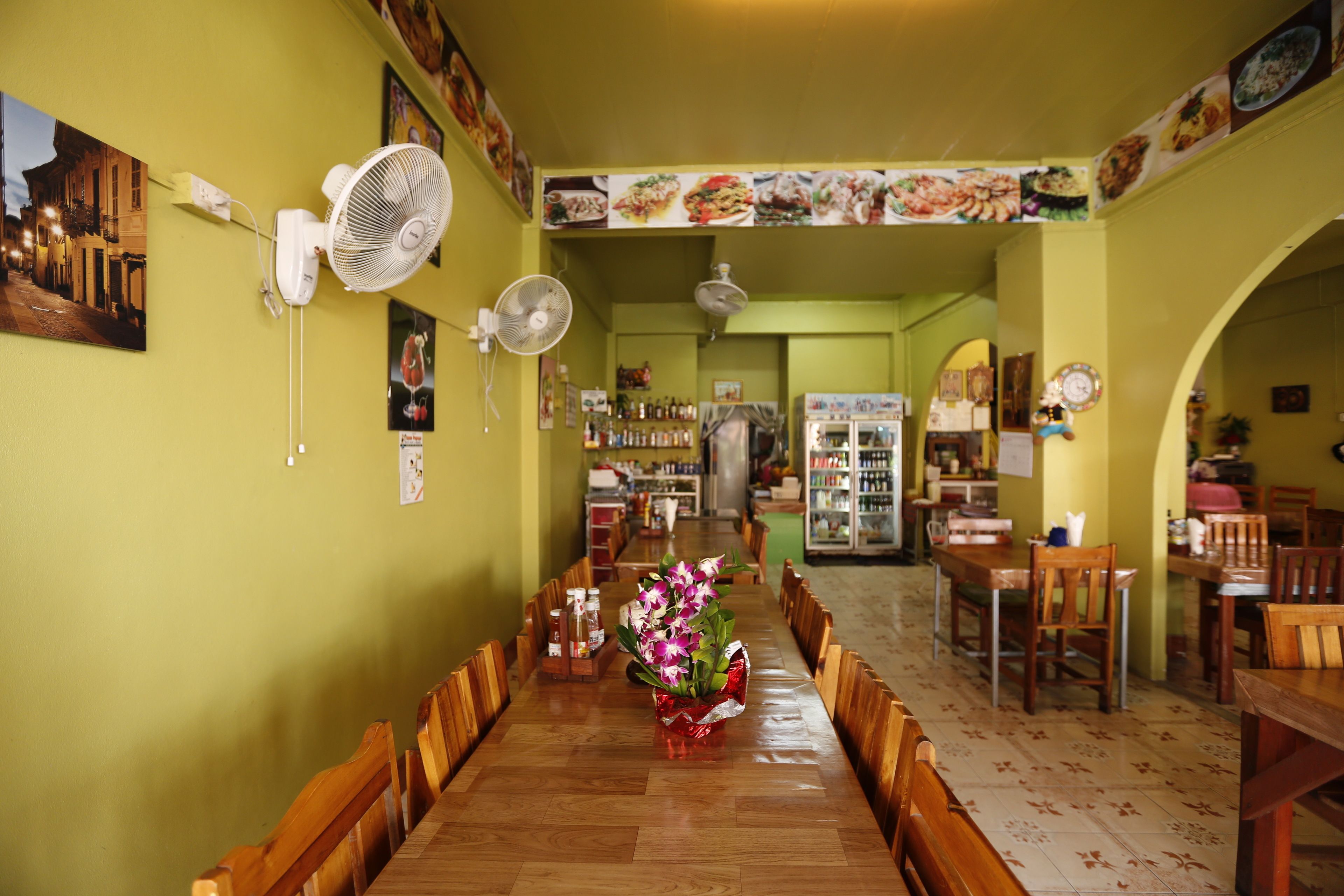 A restaurant with a long wooden table, chairs, and a vase of flowers. There are fans on the walls and a menu board above the counter.