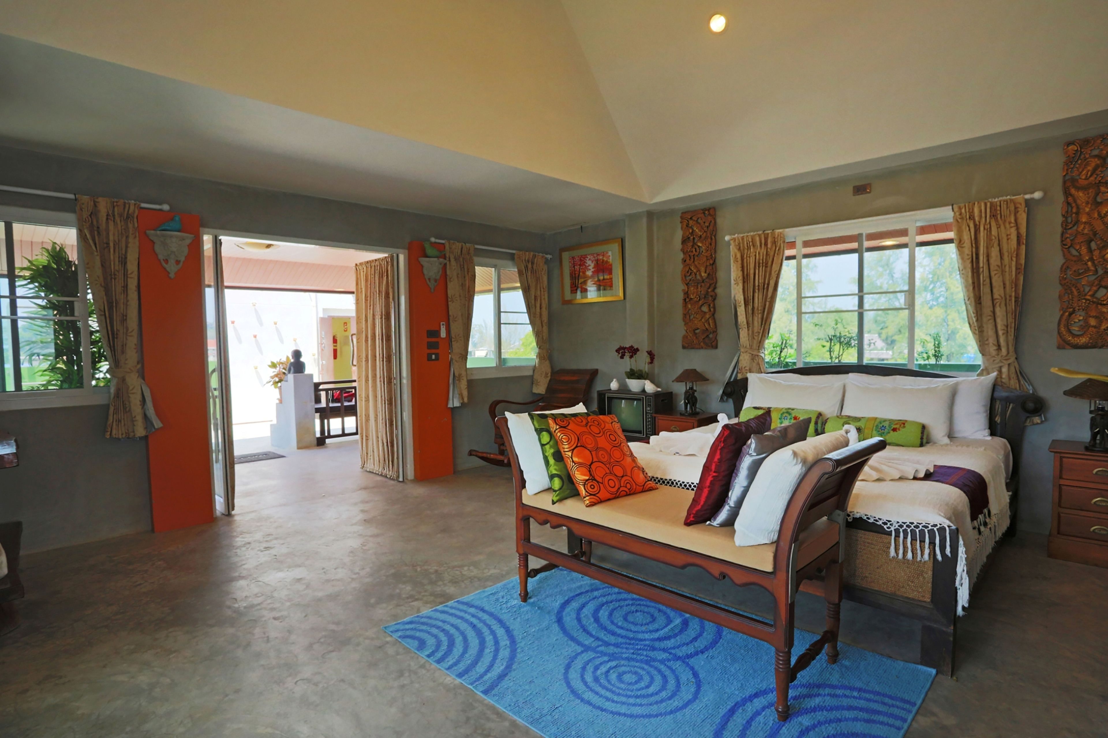 A spacious bedroom with a large bed, a wooden bench, and a desk with a chair. There are multiple windows with curtains, a view of the outdoors, and a ceiling light.