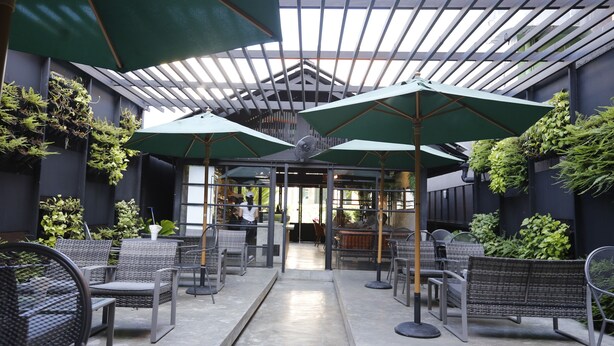 Ein Straßencafé mit Korbmöbeln, grünen Sonnenschirmen und Pflanzen vor einer dunklen Wand.