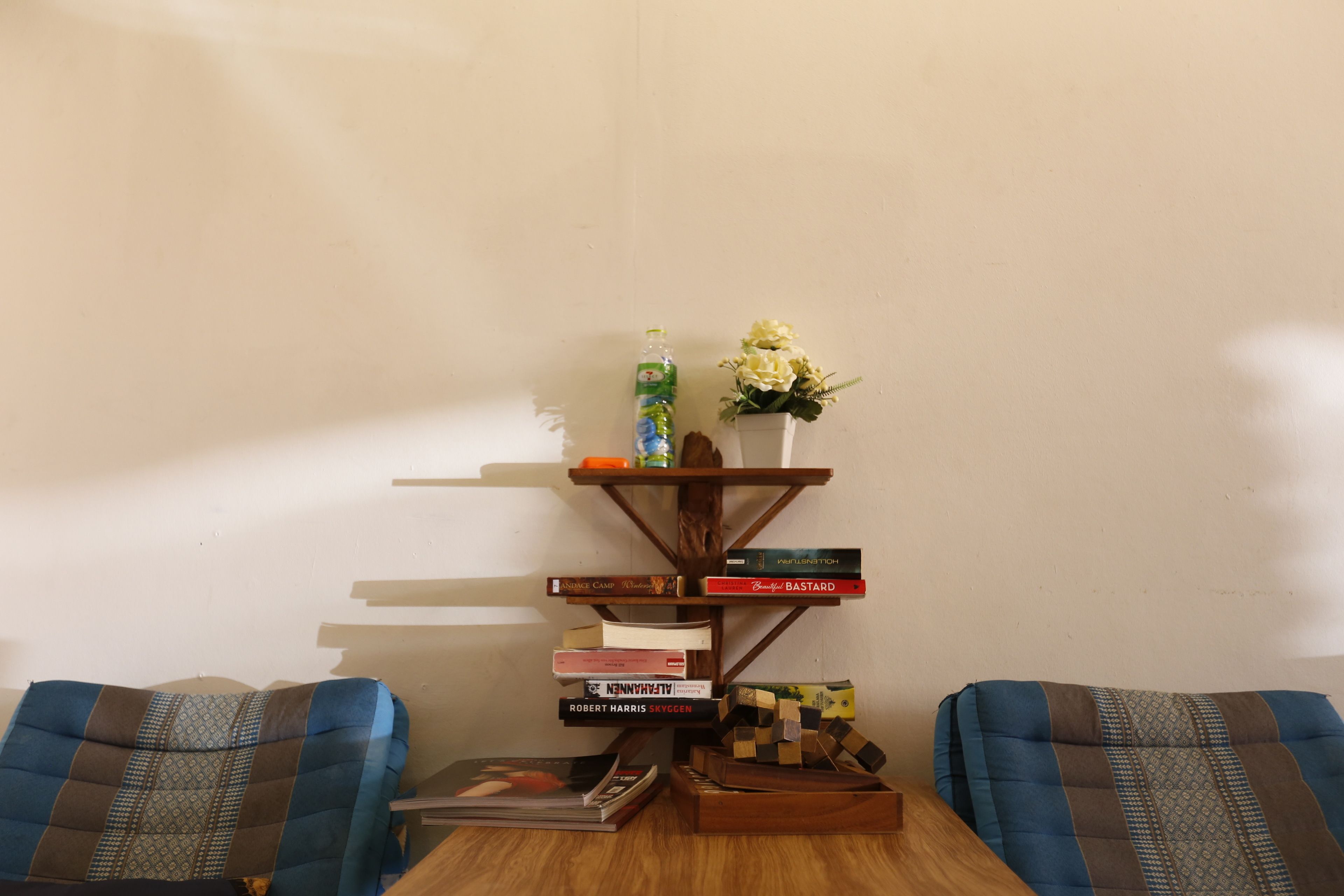 A room with a wooden table, two blue patterned chairs, a bookshelf with books and a small plant, and a wall with a shadow.